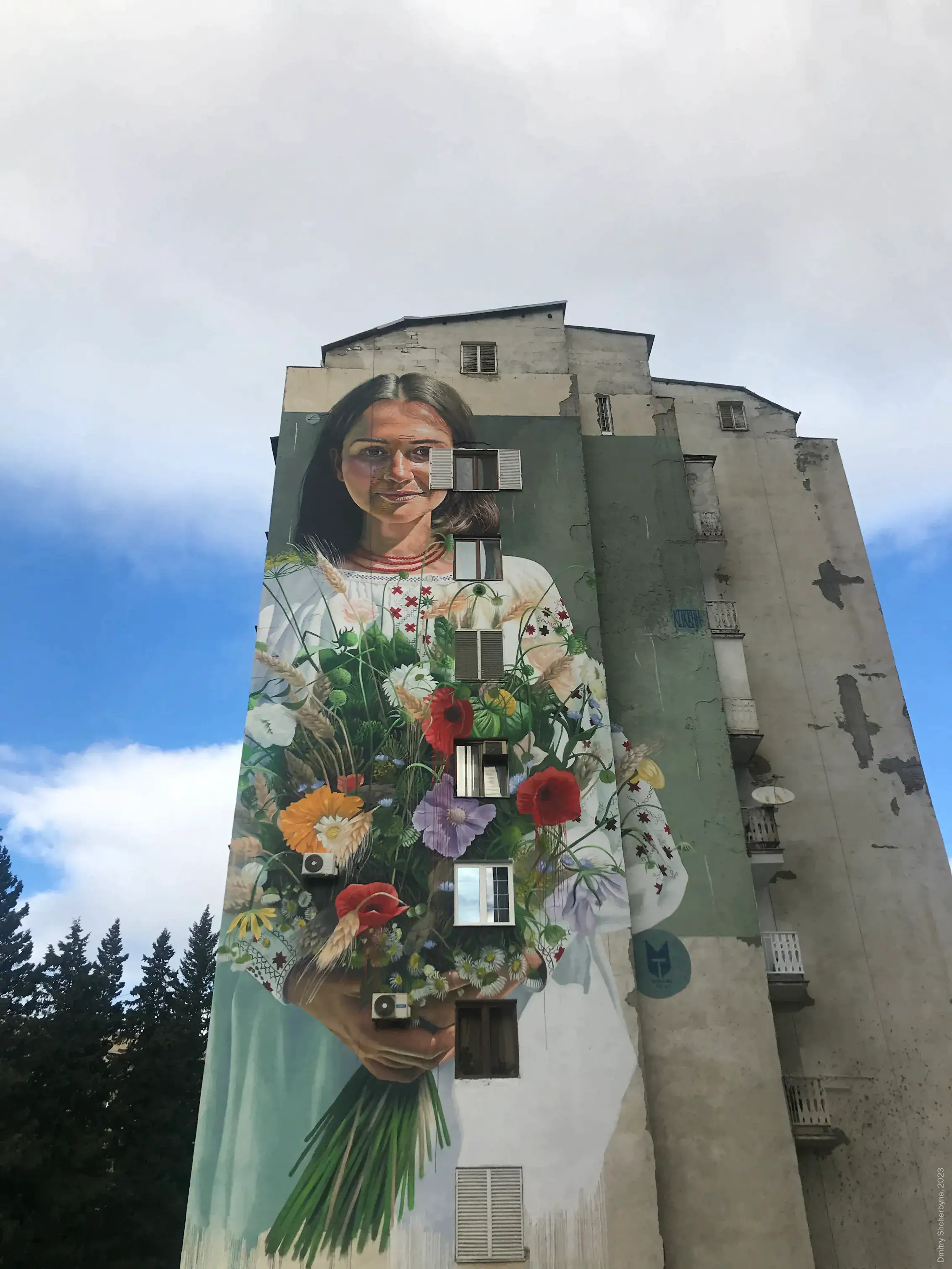 A mural spanning the full height of an apartment building depicts a woman in a traditional Ukrainian embroidered blouse, holding an overflowing bouquet of wildflowers, including poppies, wheat, daisies and irises.