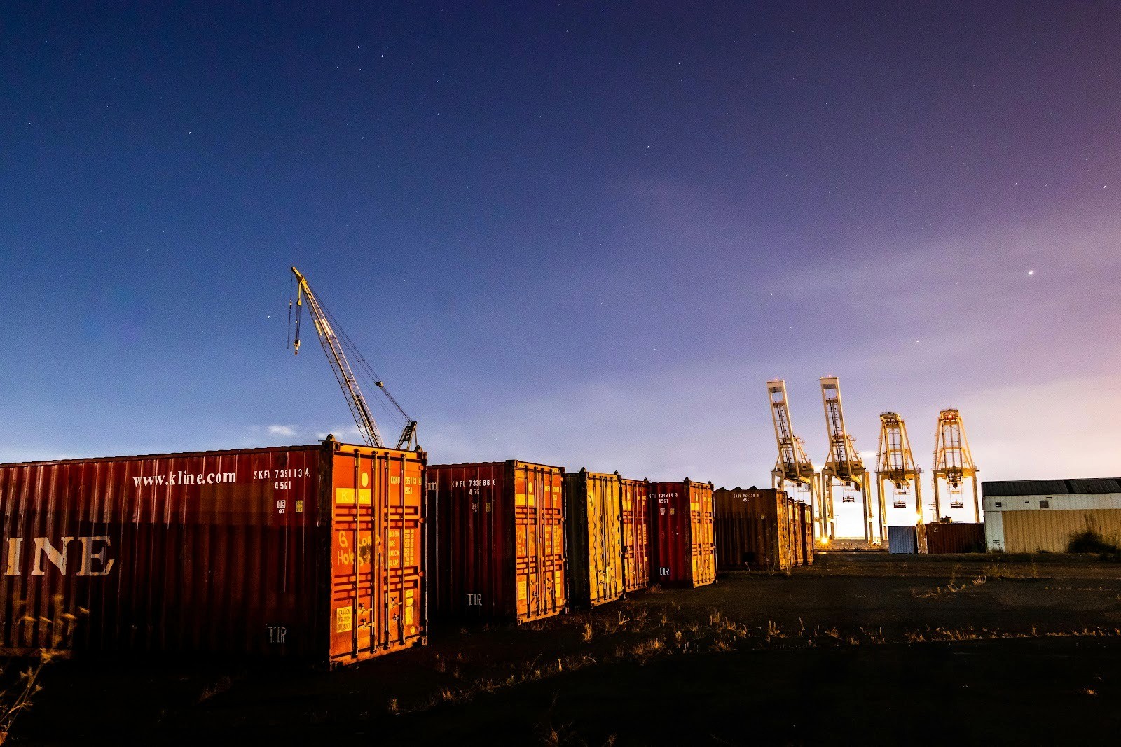 A scenic night shot of a shipping yard with cargo containers and cranes under a starry sky.