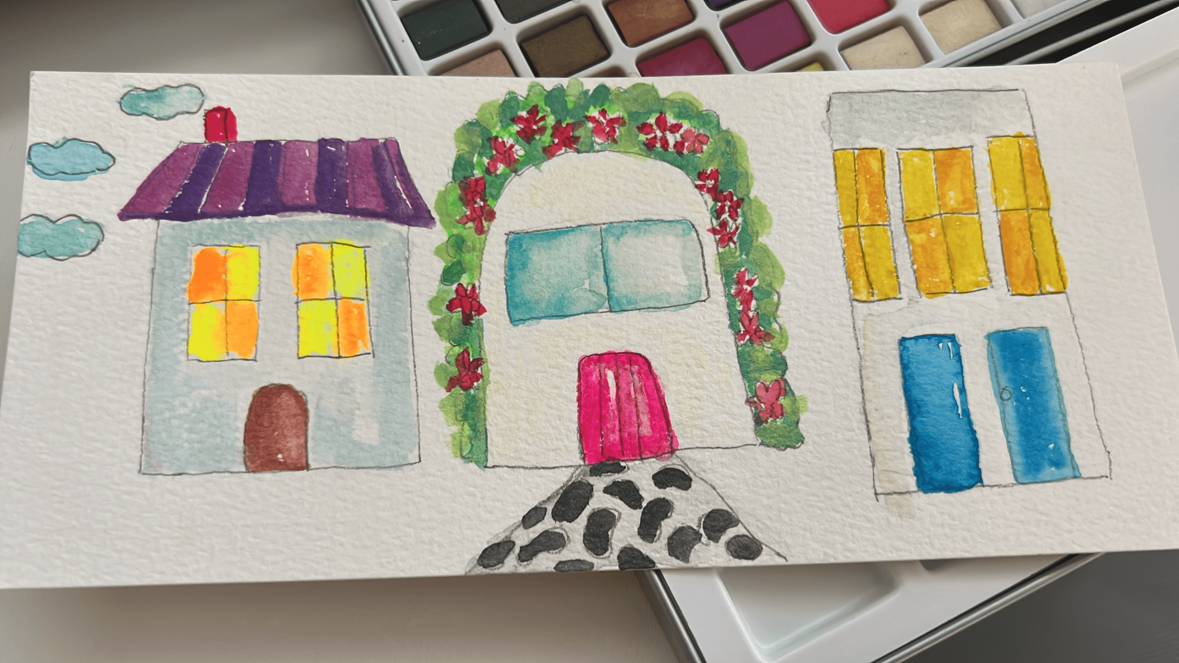 Watercolor of three little houses with lit windows, next to the paint palette