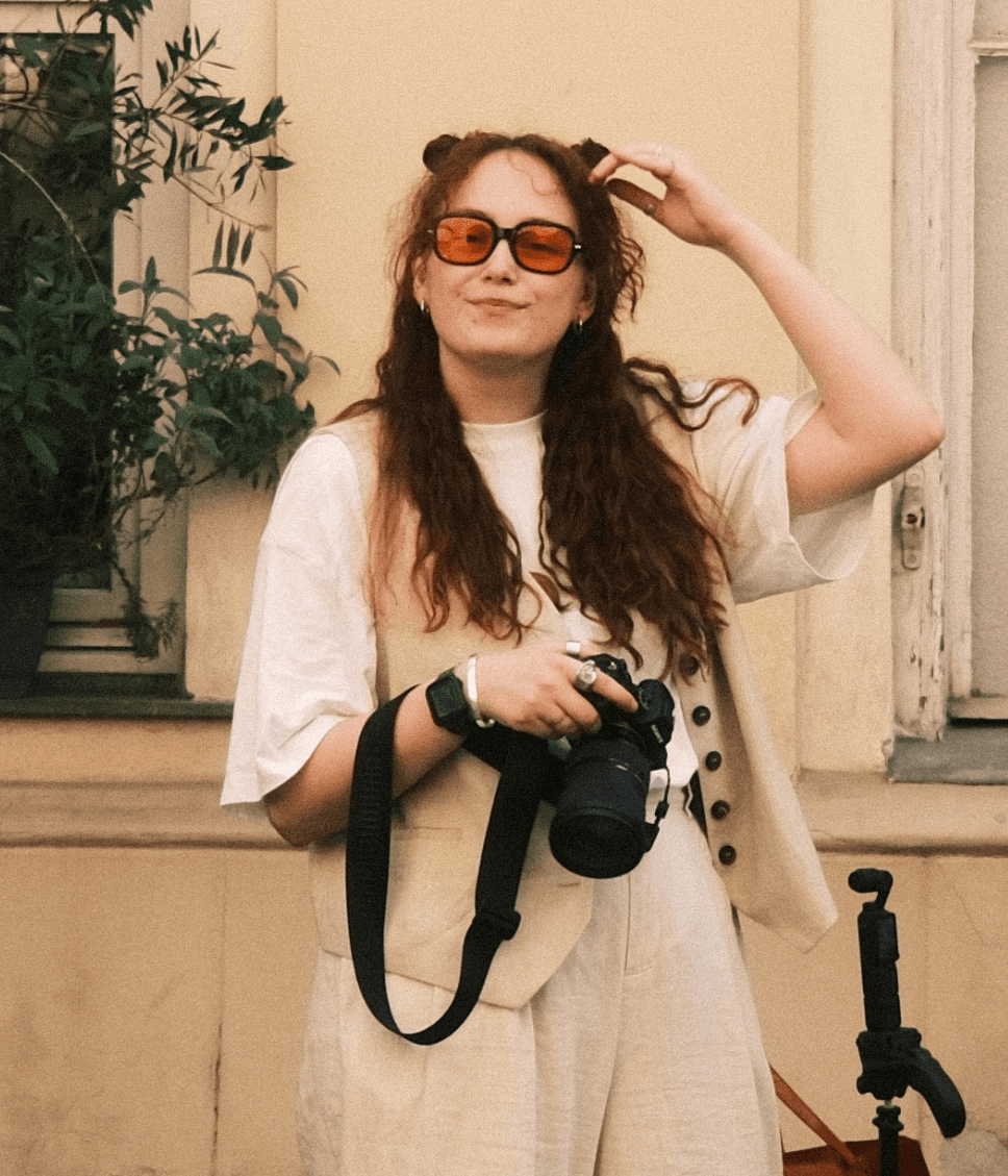 Creative director of a digital agency, wearing neutral-toned overalls and tinted sunglasses, holding a professional camera. Standing outdoors with a relaxed yet confident vibe, she represents the agency’s visual storytelling and content creation expertise.