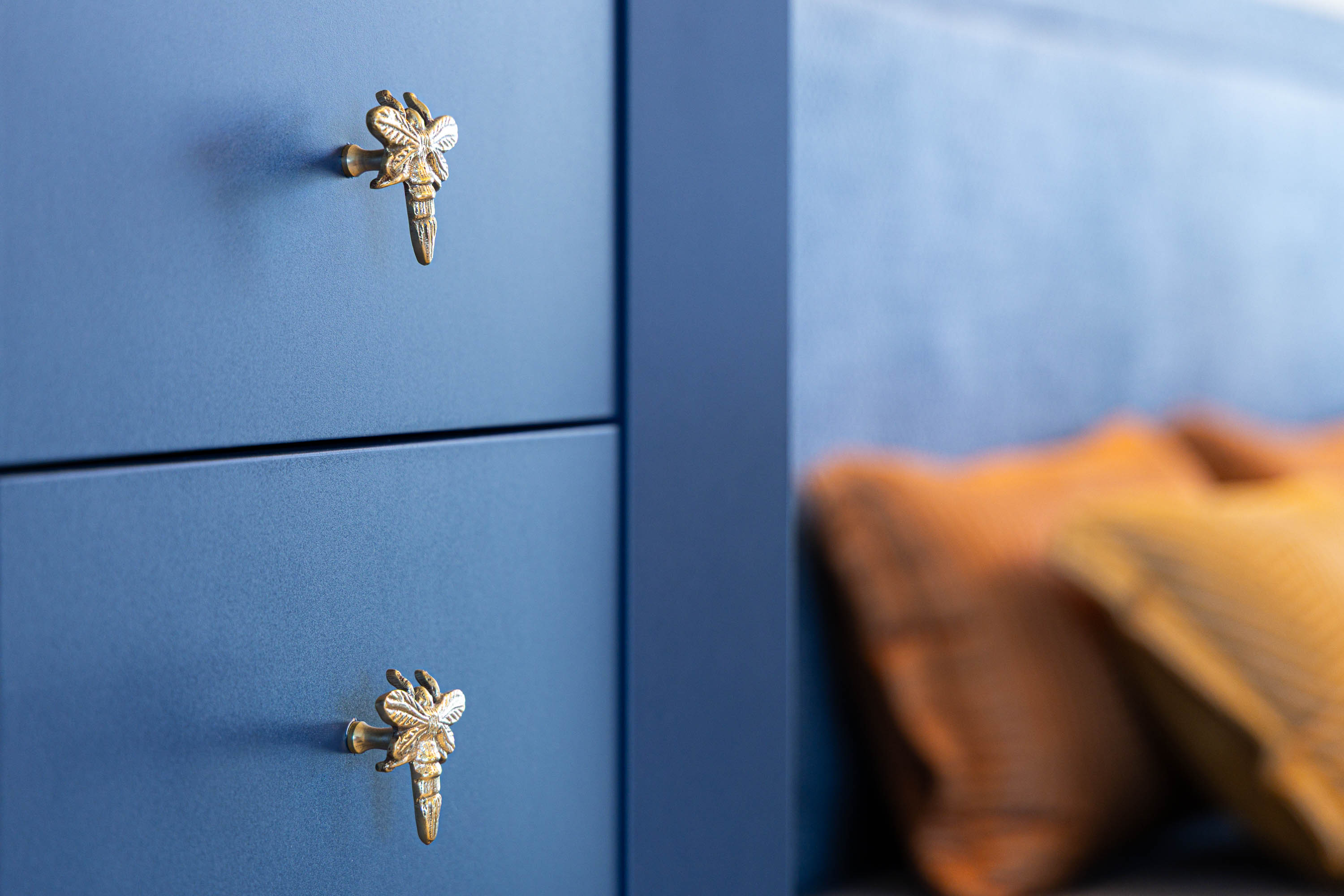 Blue chest of drawers with brass dragonfly handles, beside a mustard cushion