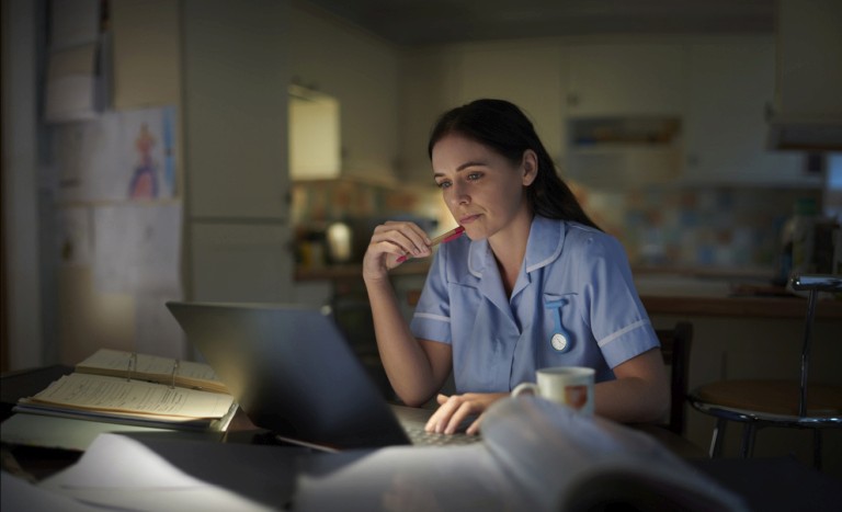 A nurse in blue scrubs sitting at a desk in a dimly lit setting, looking at a computer screen during what appears to be a night shift