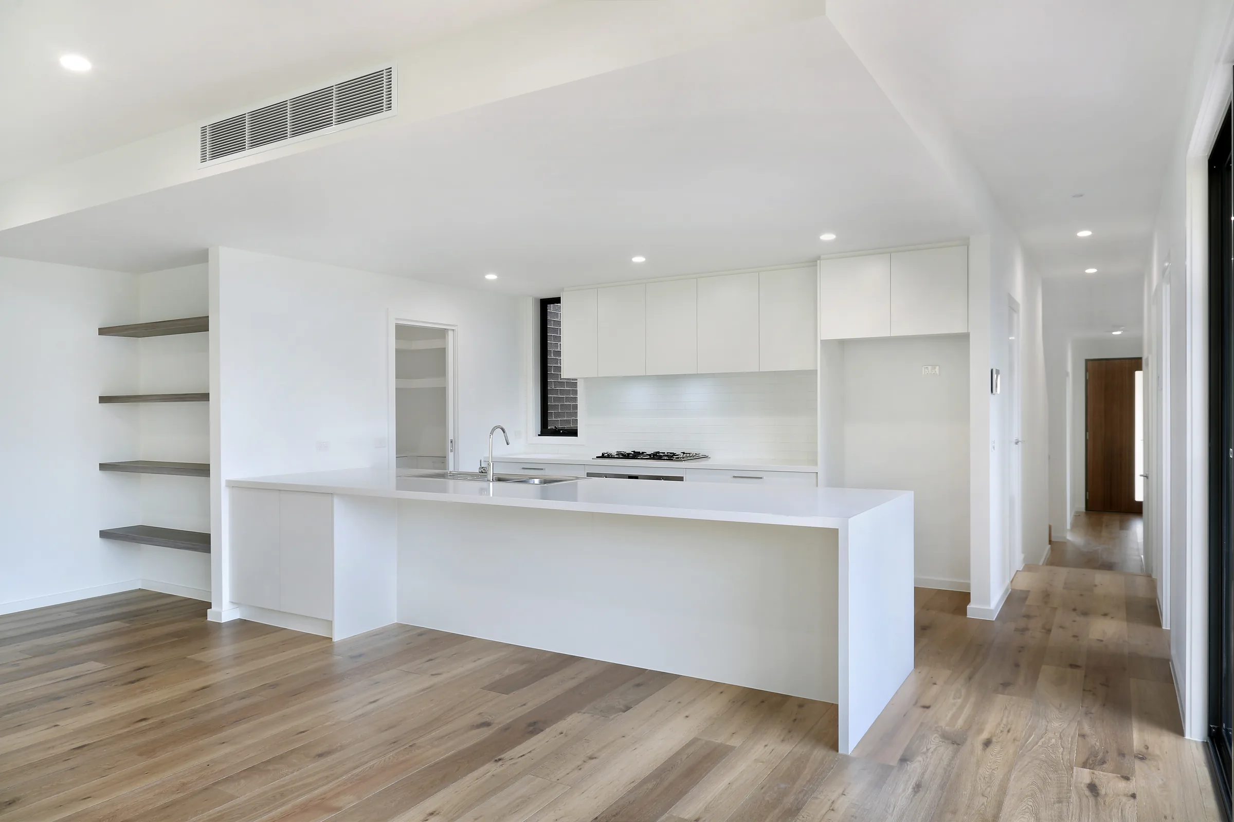 Open-plan kitchen and living with white cabinetry, a white waterfall island, grey floating shelves, gas cooktop, chrome tap and oak floors, with a hallway leading to a timber front door
