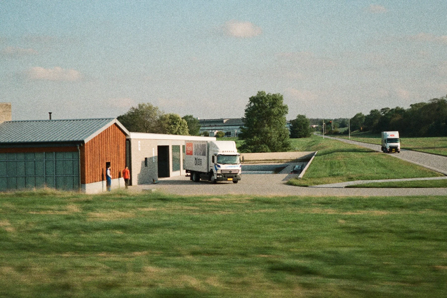 Logistiek bedrijfsterrein met een kantoorgebouw, laadperron en twee vrachtwagens in ochtendlicht.