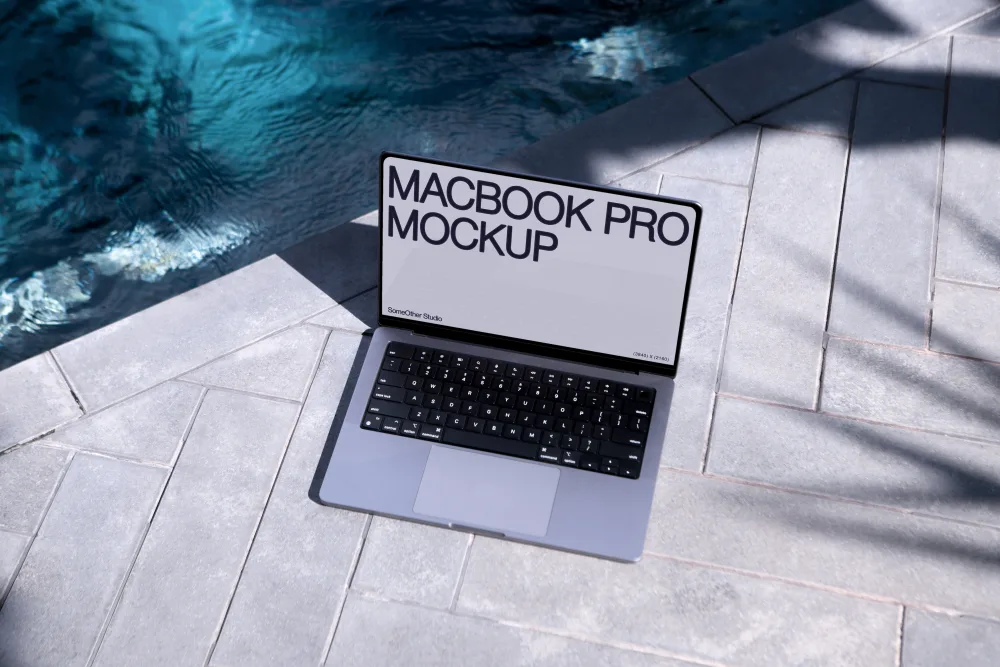 A MacBook Pro mockup placed on gray tiles beside a pool with shimmering blue water, surrounded by soft shadows from nearby foliage in bright sunlight, creating a relaxed outdoor setting.