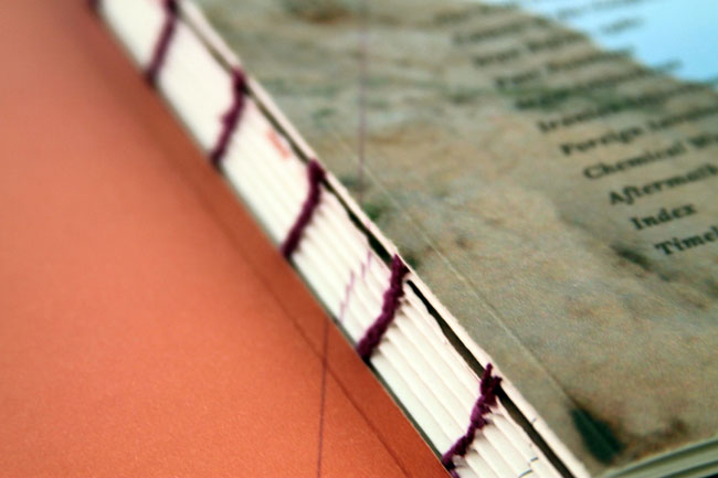 A close-up of a book with a white cover and off-white pages, bound by dark purple string. The pages are slightly fanned out, and the cover rests on an orange surface. A blurred map or chart is visible in the background, with text including “Foreign Language,” “Chemical,” “Aftermath,” and “Index Time” partially visible.