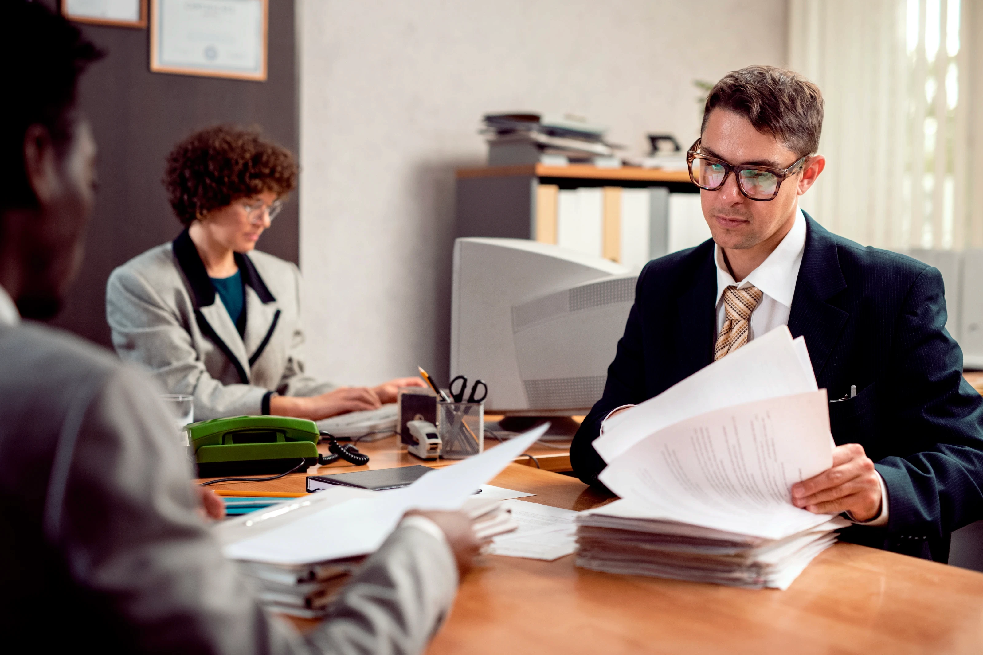 A man in a suit reviews documents at a desk, focused and professional. In the background, a woman works on a computer. The setting is an office.