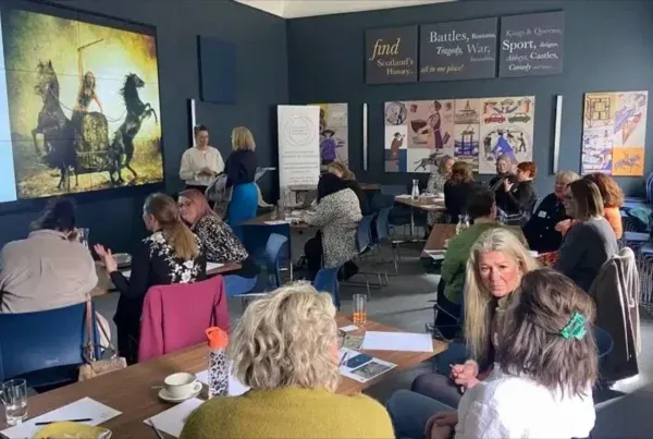 A group of people in a workshop setting with a presentation of knights on horses. Attendees sit at tables, engaged in discussions, under a historical-themed decor.