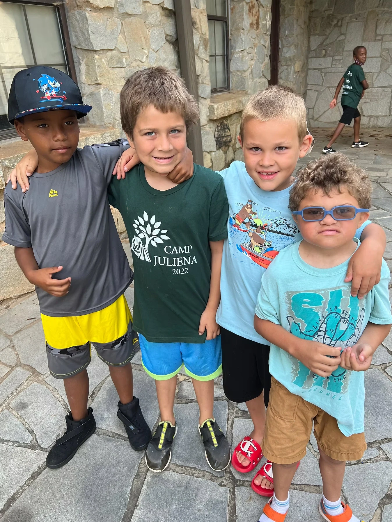 Four boys at Camp Juliena stand together, smiling for a photo. One is wearing a Camp Juliena shirt.