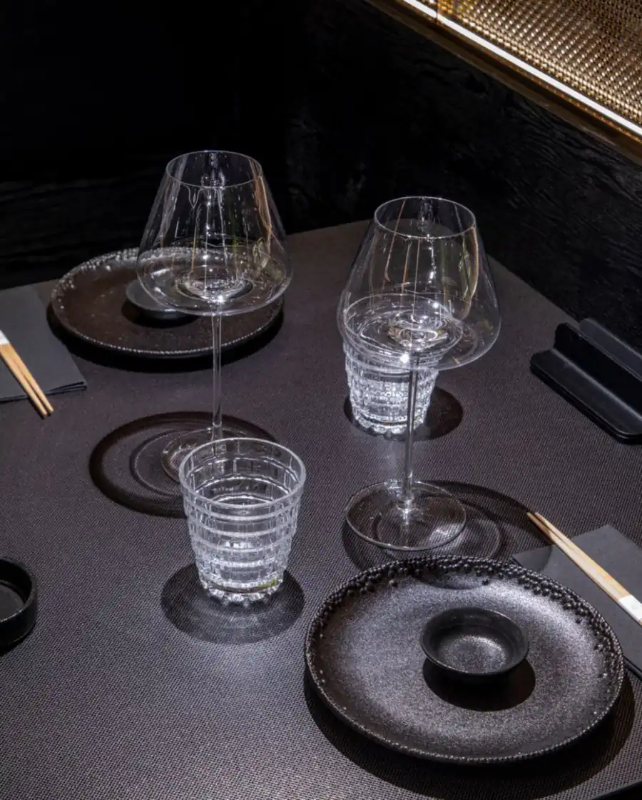 Table dressée de verres en cristal, assiettes noires et baguettes, sous une paroi de mailles de cuivre