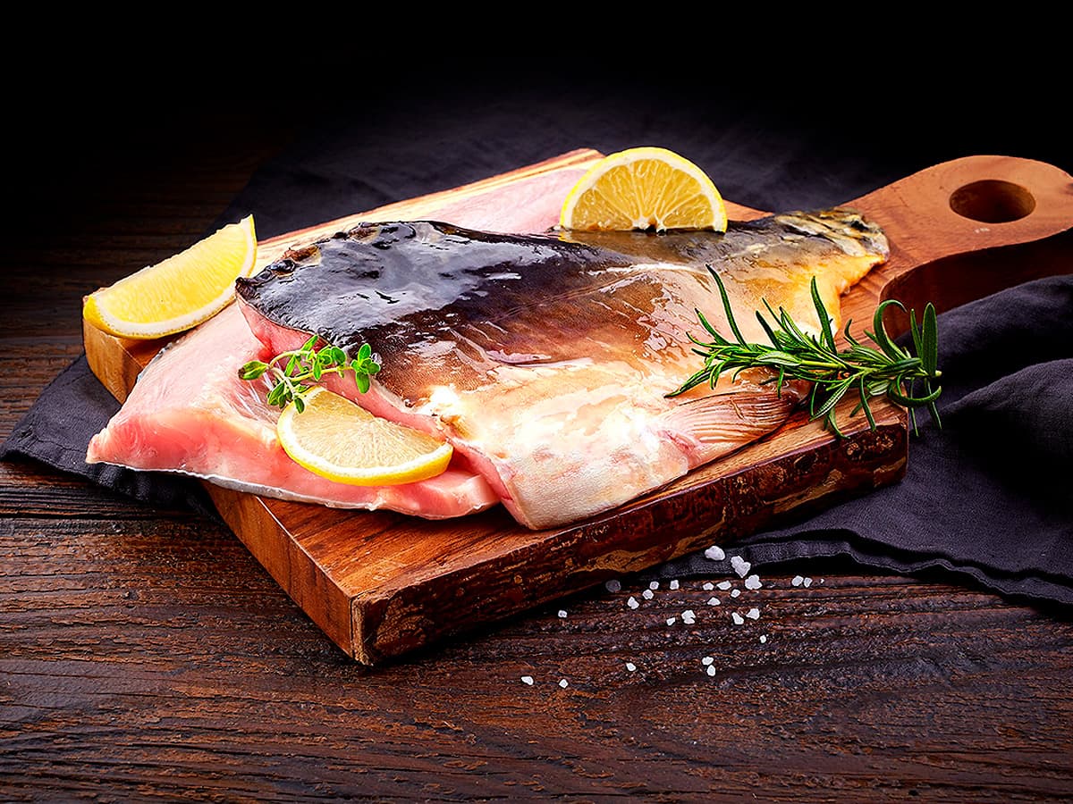 A raw fish carp fillet is displayed on a wooden cutting board, garnished with lemon wedges and fresh rosemary sprigs, set against a dark cloth and wooden table for a rustic presentation.