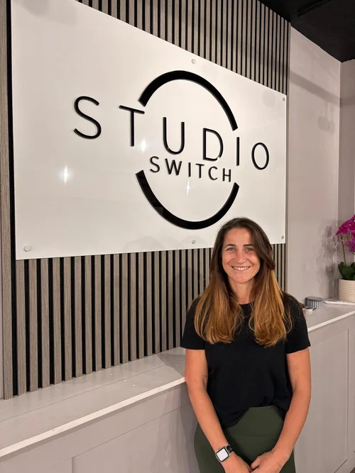 Frankee, a Studio Switch Pilates instructor, seated beneath the Studio Switch sign at the reception area.
