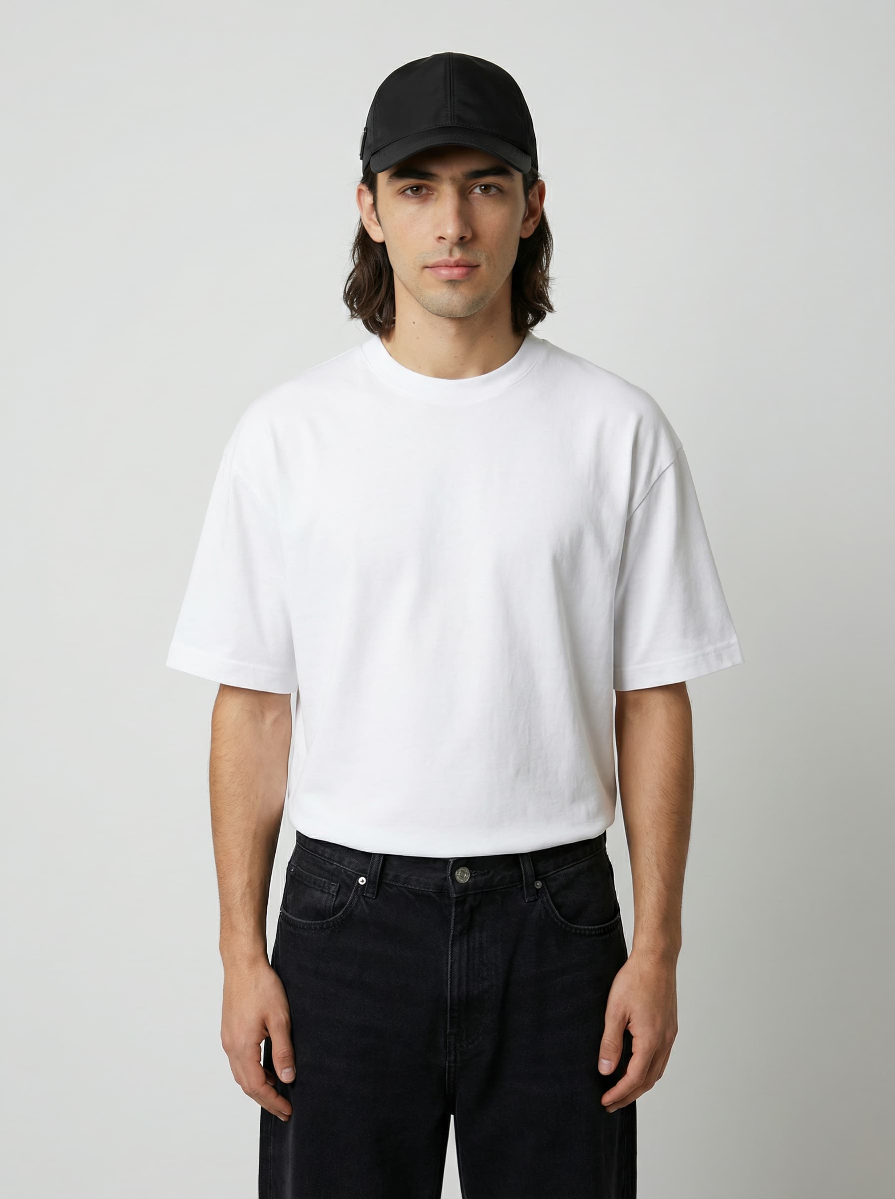 A person wearing a plain white t-shirt and a black cap stands against a neutral gray background, looking directly at the camera, with their hands relaxed by their sides.