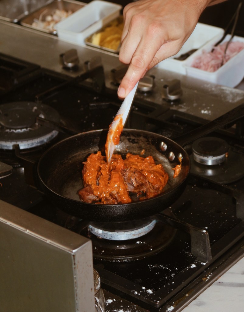 A chef's hand holding a spatula, stirring a savory orange sauce with pieces of meat in a pan on a stovetop, surrounded by various ingredients in containers.