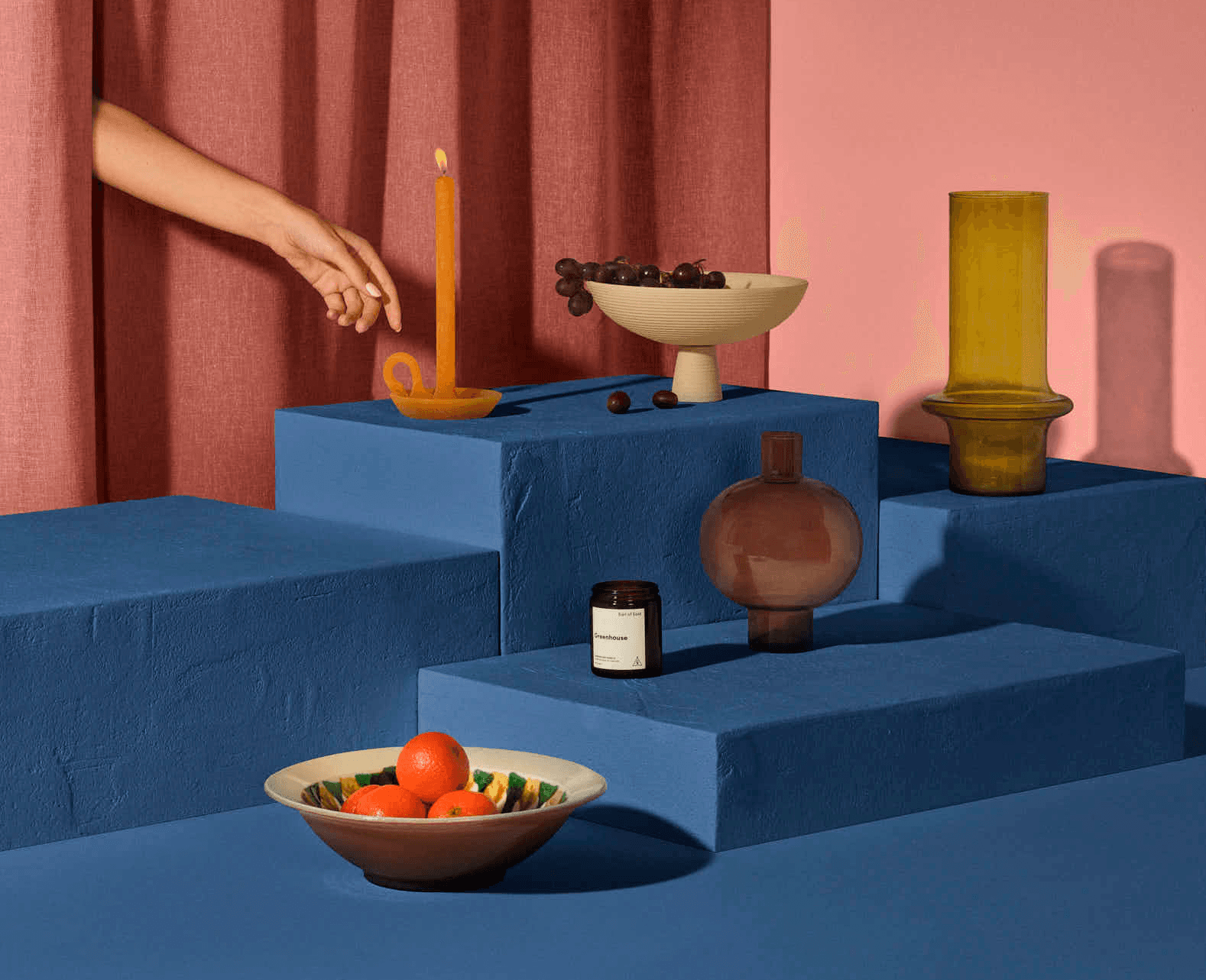 A vibrant still life featuring various ceramic vessels and fruits on blue platforms, with a hand reaching towards them.