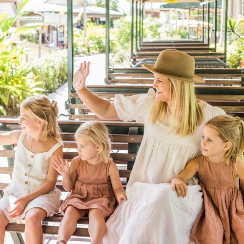 Seorang wanita dan tiga gadis muda melambaikan tangan sambil duduk di trem terbuka berwarna hijau di lingkungan yang subur dan cerah.