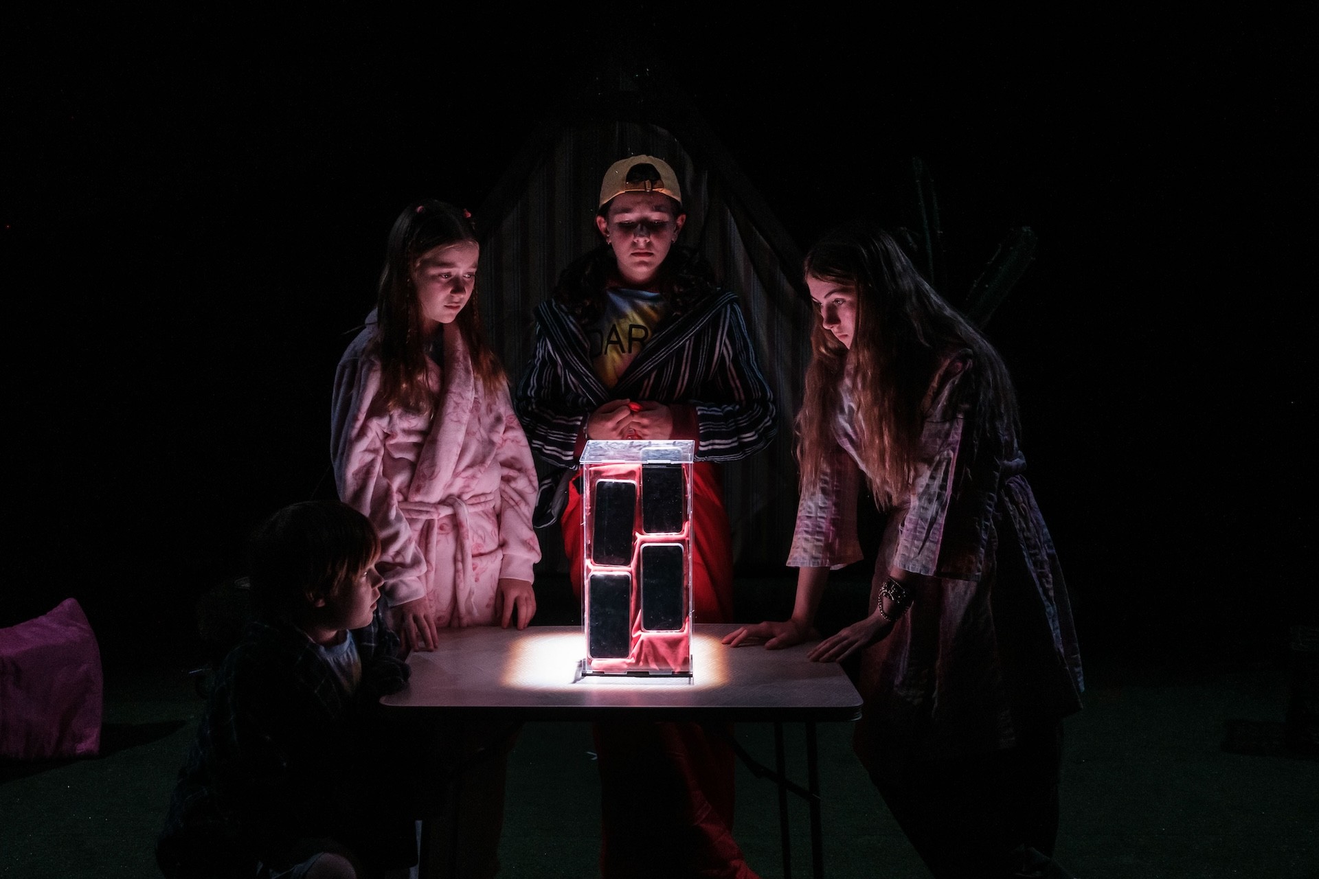 Four children in pyjamas and robes gather around a glowing tower of confiscated mobile phones on a table in the dark, lit dramatically from below as they stare at the illuminated structure.