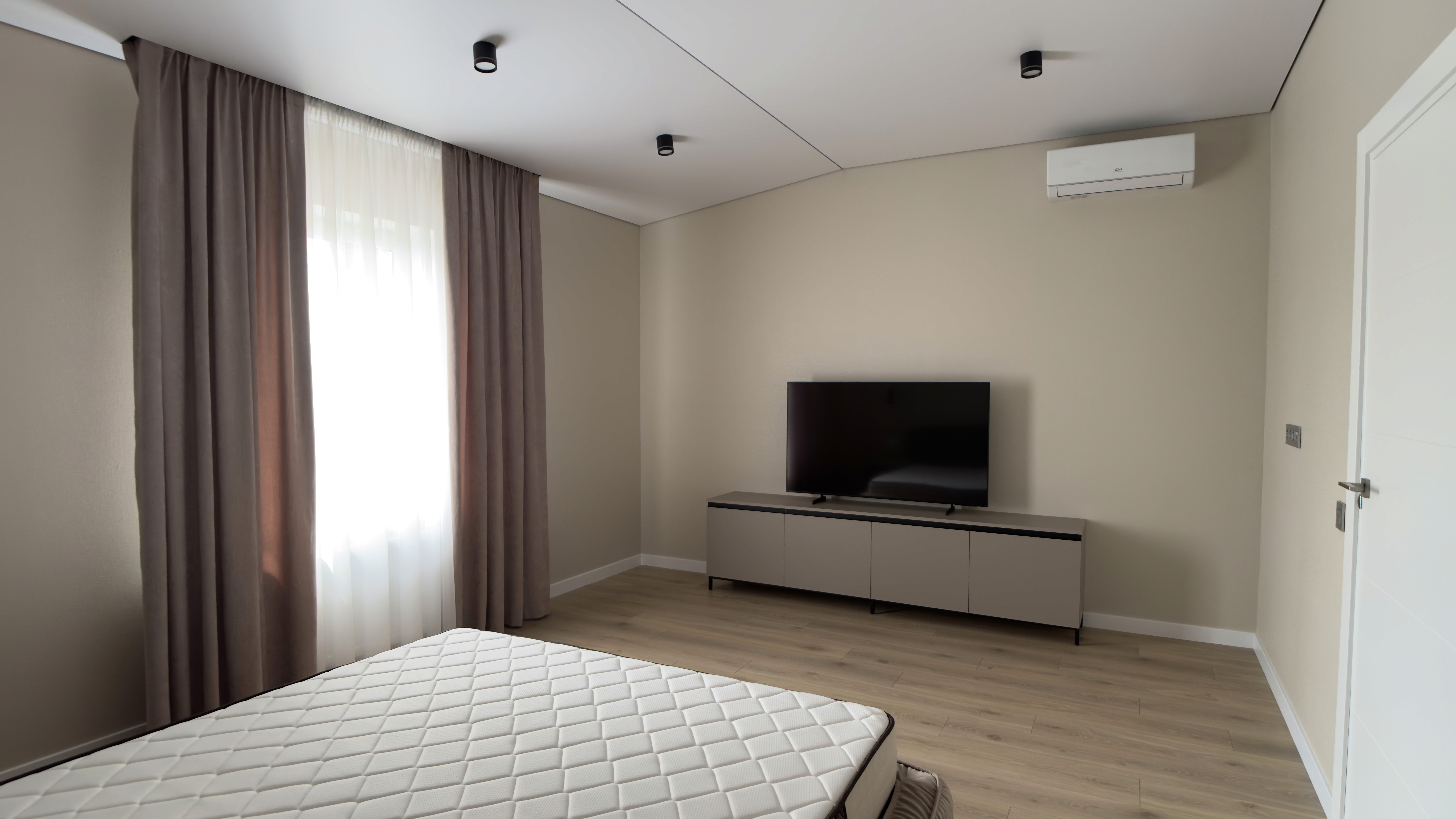Світла спальня з телевізором на тумбі, кондиціонером і великим вікном із щільними шторами