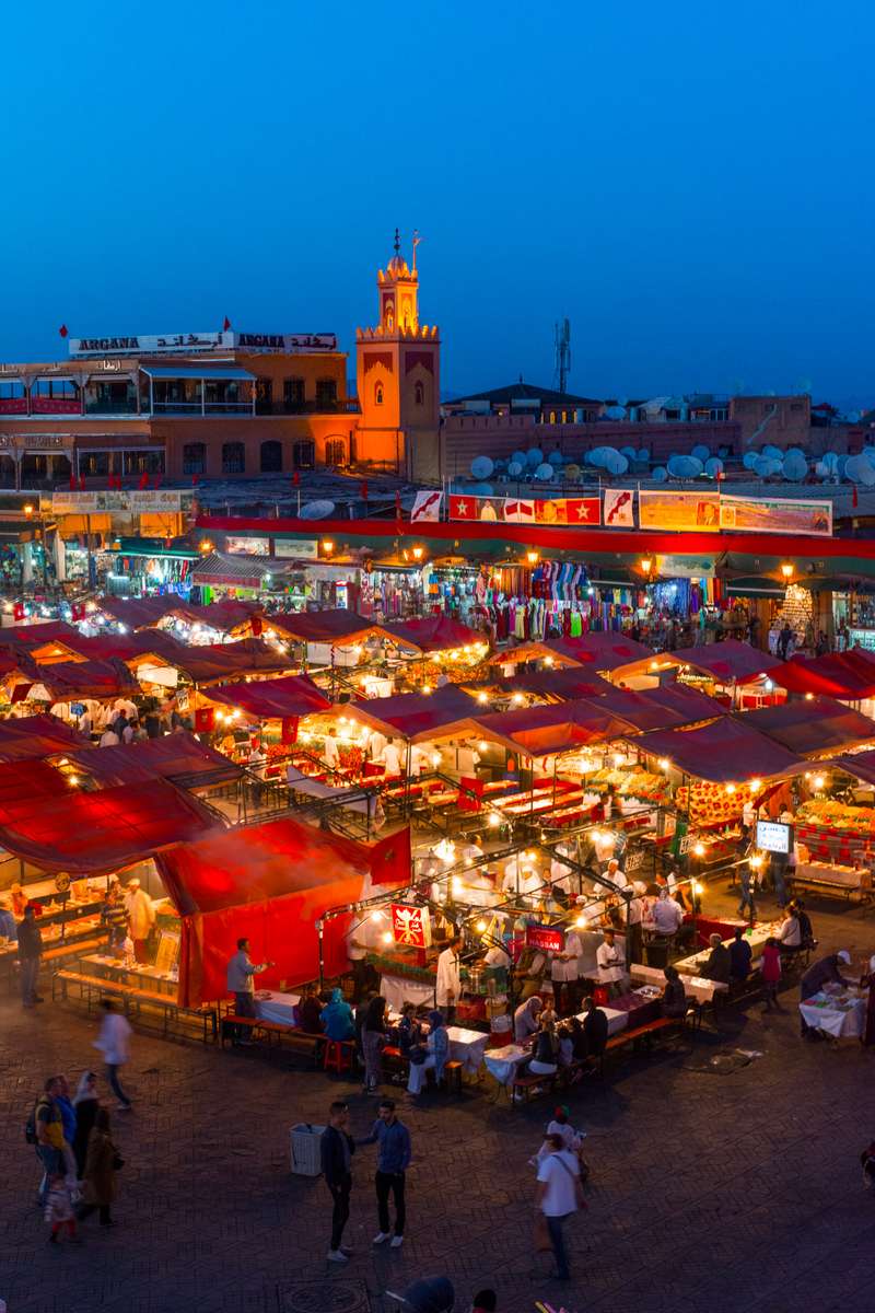 A crowded beach in the evening with red and yellow umbrellas, lit stalls, and a tall clock tower under a blue sky.