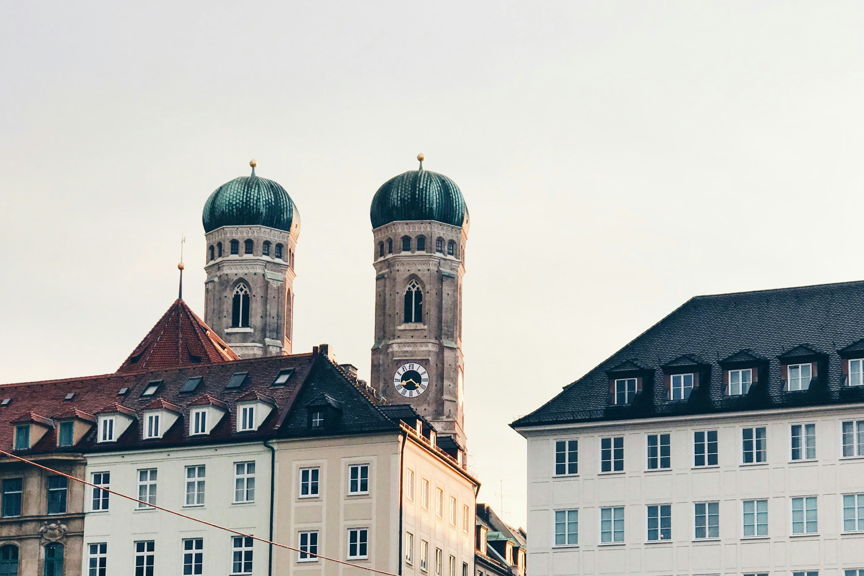 A view of the iconic twin domes of the Frauenkirche cathedral rising above a row of classic Bavarian architecture buildings in Munich, Germany, under a clear sky.