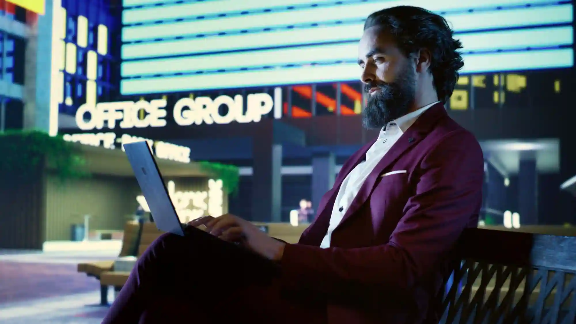 A businessman in a suit working on his laptop outdoors at night near an illuminated office building.