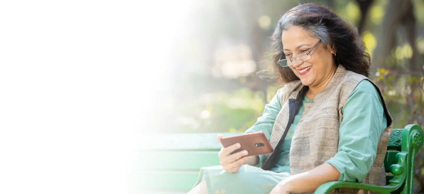 An older Indian origin woman sitting on a park bench, smiling while using her smartphone, representing the ease of access to digital health solutions for older adults managing chronic conditions like diabetes.