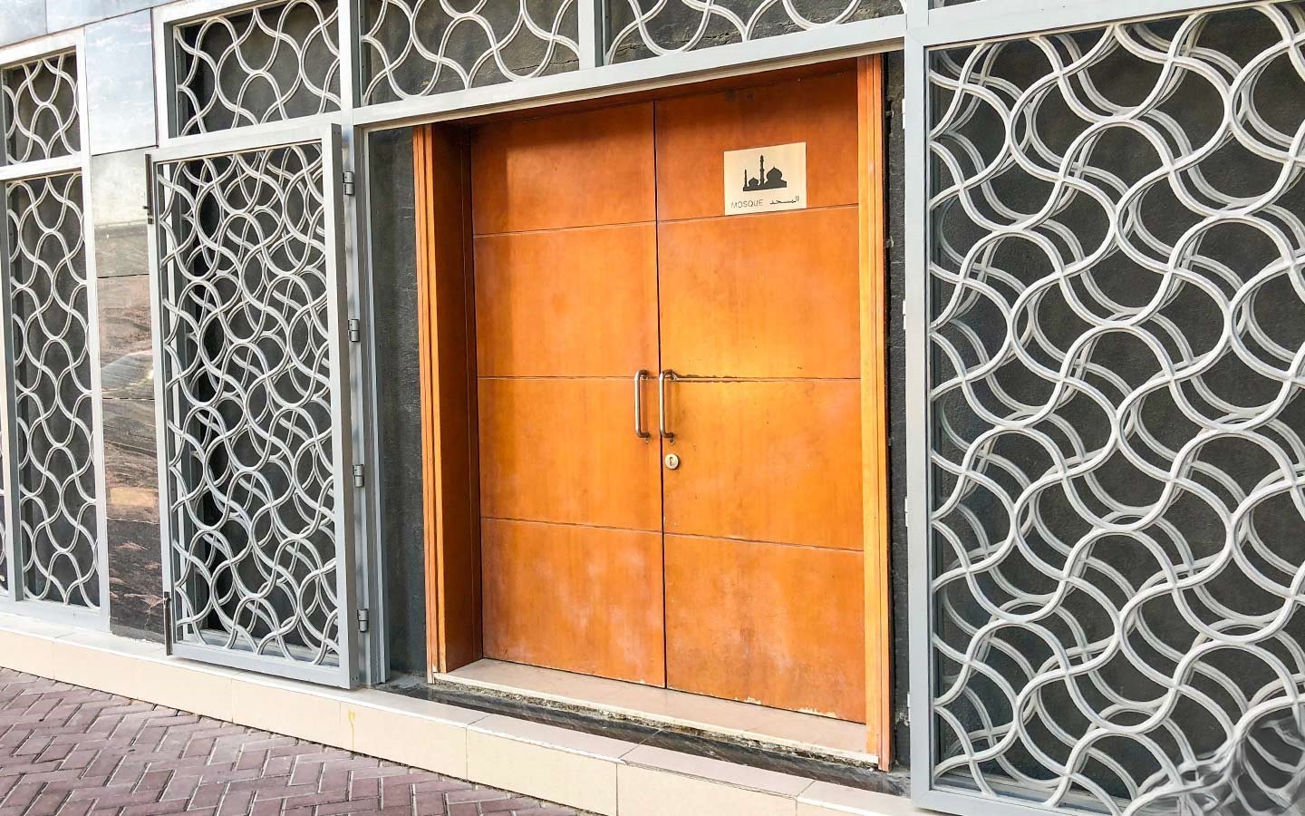 Jumeirah Lake Towers mosque entrance with a wooden double door and white geometric, wavy patterned screens on either side.