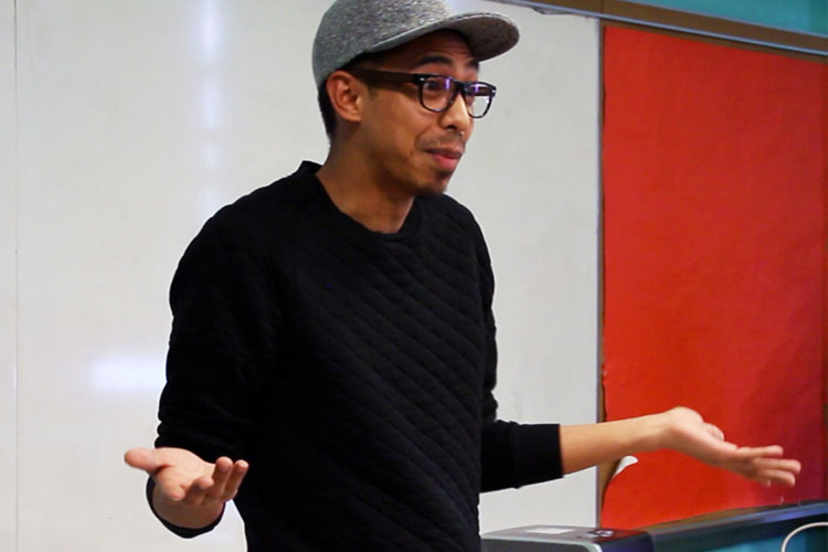 A man wearing a black long-sleeved shirt and a gray baseball cap stands in front of a white wall and a red chalkboard. He wears glasses and has his arms slightly raised and hands open, as if gesturing while speaking or explaining something. The chalkboard is positioned to his right, and the white wall extends to the left. A portion of a gray object is visible in the bottom right corner of the image.