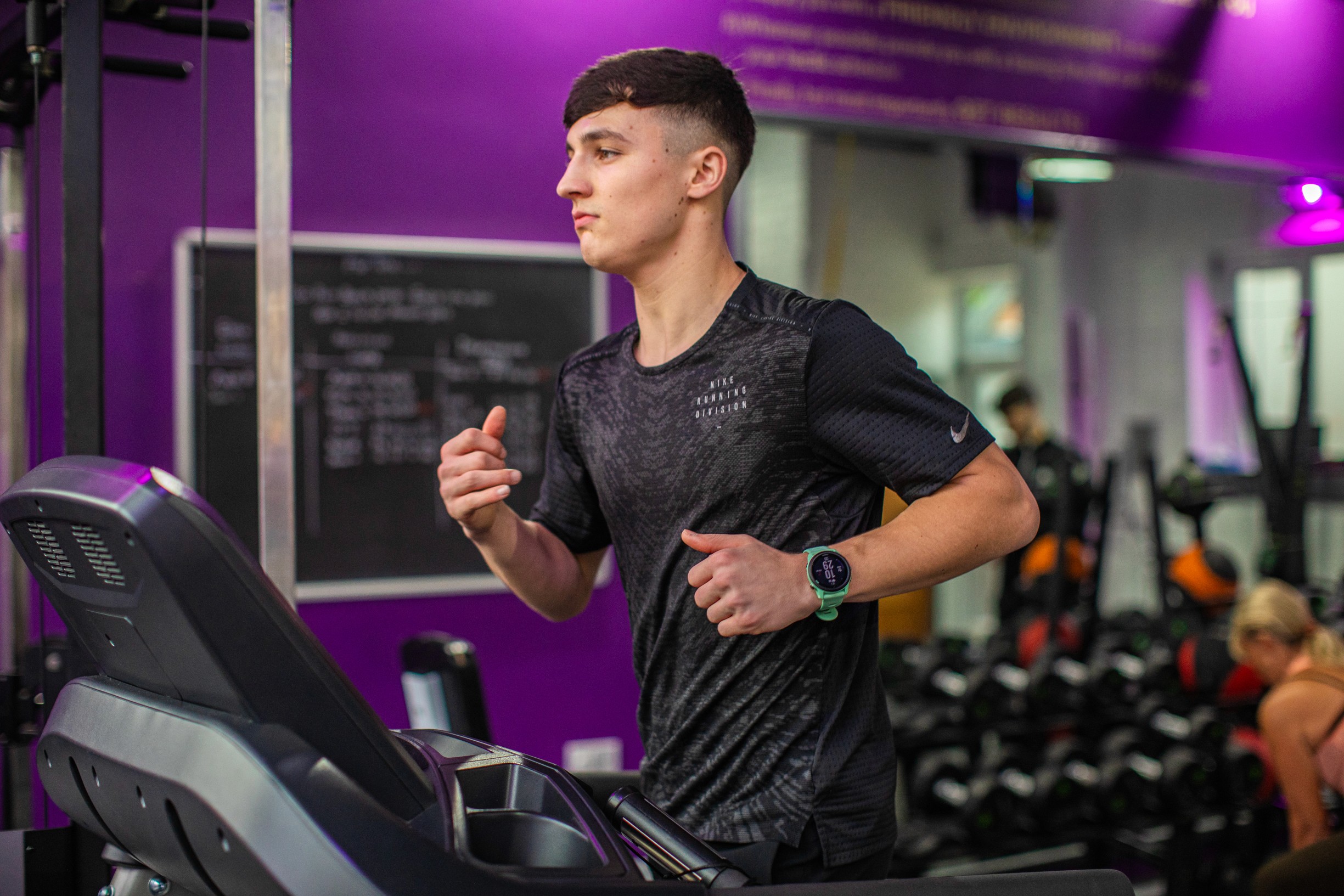A person jogs on a treadmill in a gym, wearing a black athletic shirt and a smartwatch, with exercise equipment in the background.