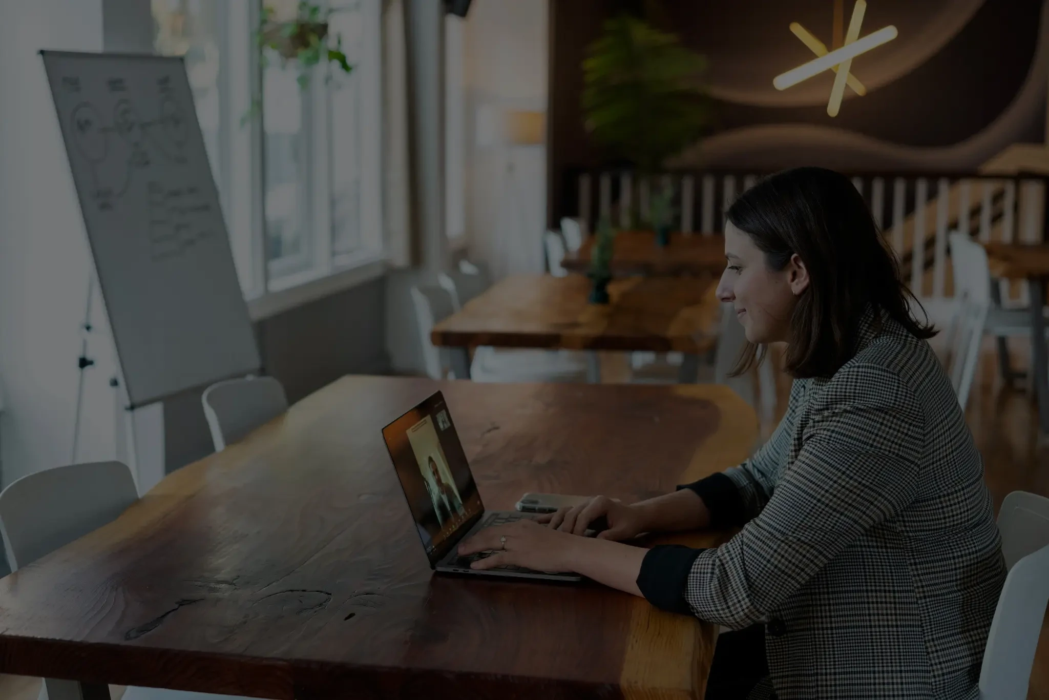 A woman works on a laptop at a large wooden table in a bright meeting room, with a whiteboard, chairs, plants, and a staircase in the background.
