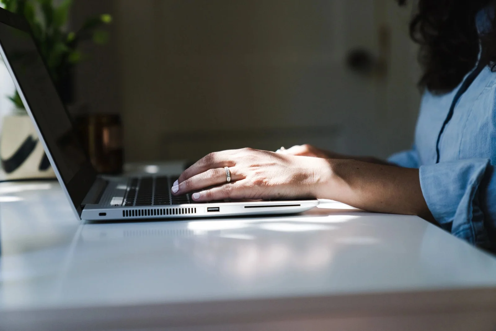A person is typing on a laptop placed on a white desk, with natural light streaming in, suggesting a study or work setting, potentially related to UCAM University.