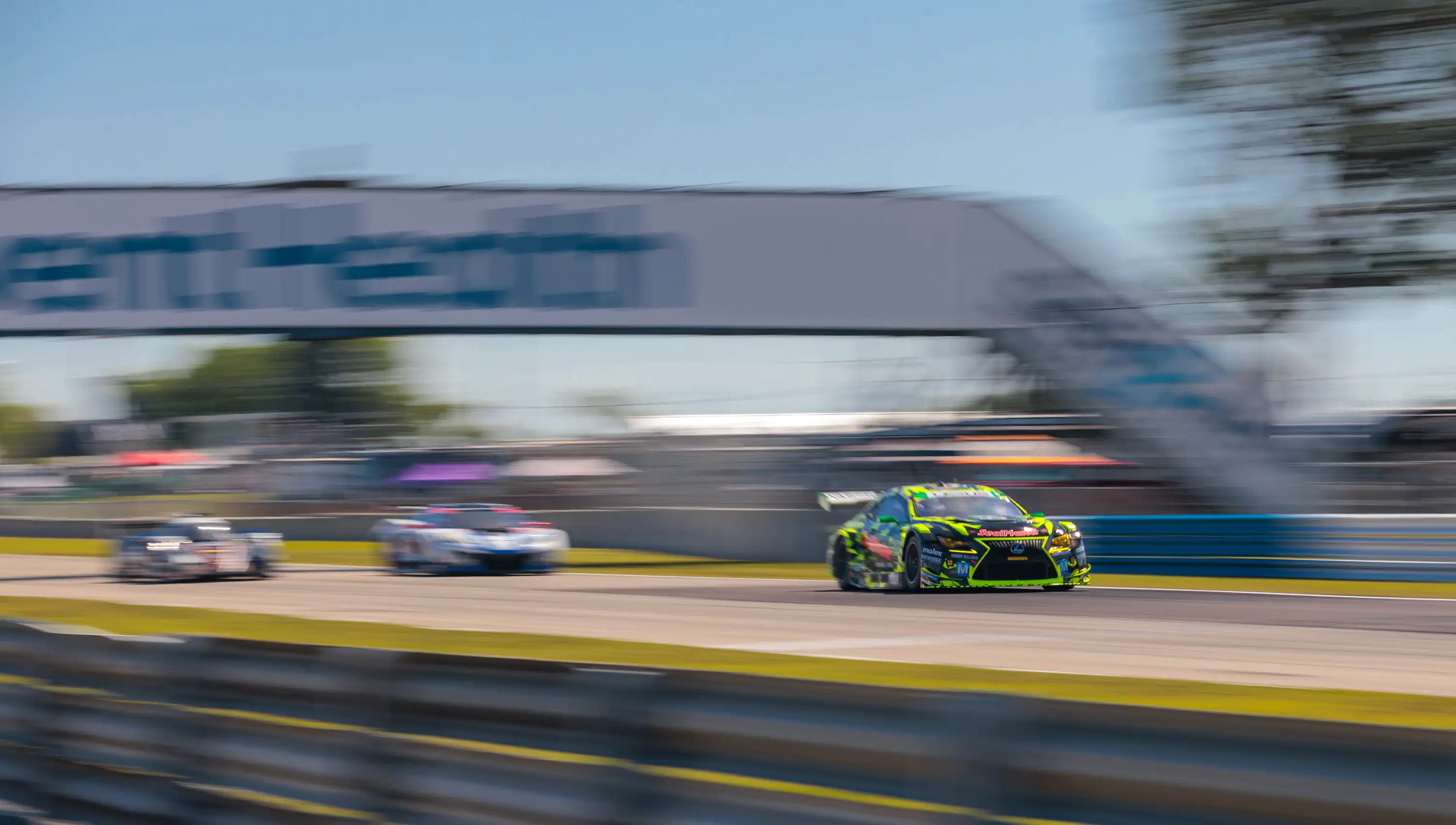 A green and yellow race car speeds on a track, closely followed by a white car, with a blurred banner and trees in the background.