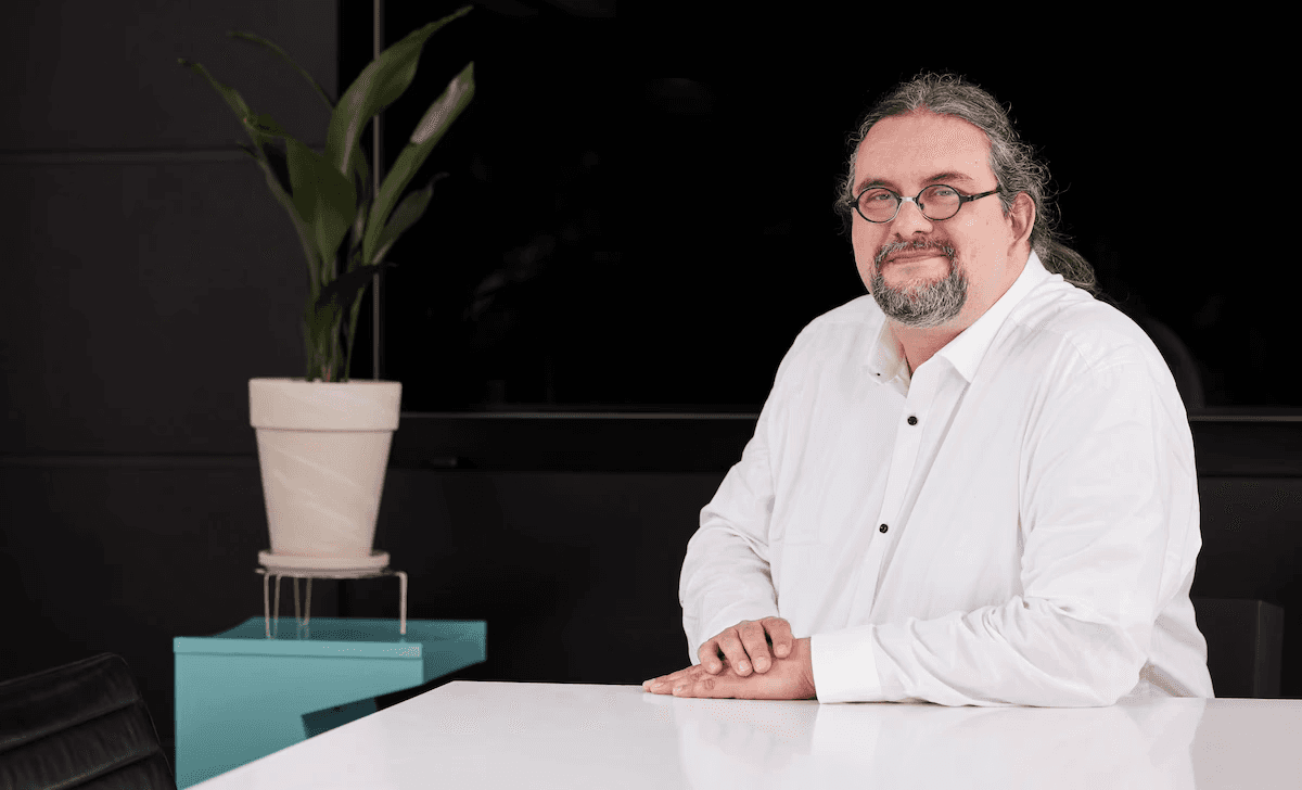 Person mit Brille und langen Haaren, zum Zopf gebunden, trägt ein weißes Hemd und sitzt im Büro an einem Tisch neben einer Pflanze