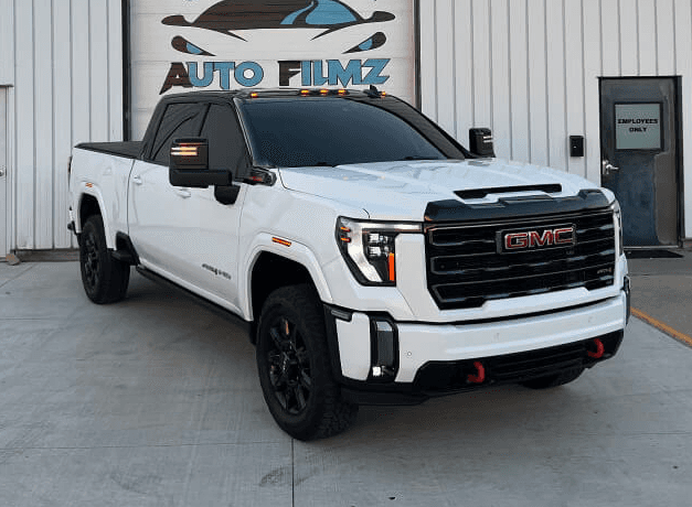 A white GMC Sierra HD AT4 pickup truck is parked on a concrete driveway in front of a white industrial building.