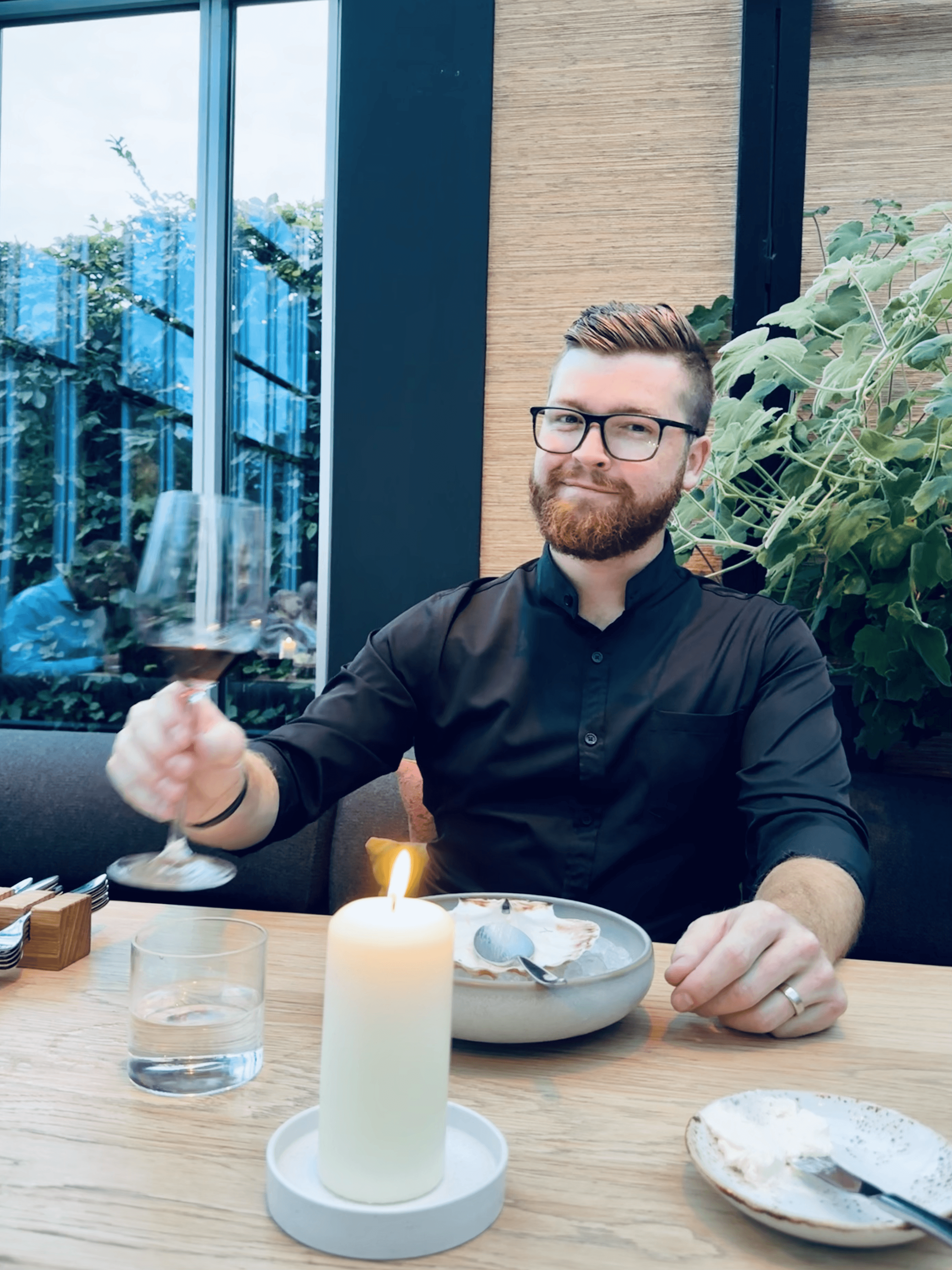 A bearded man (named Mitch) wearing glasses and a black shirt is smiling while holding a glass of red wine, sitting at a wooden table with a lit candle, a glass of water, and a bowl, set against a backdrop of large windows and greenery.