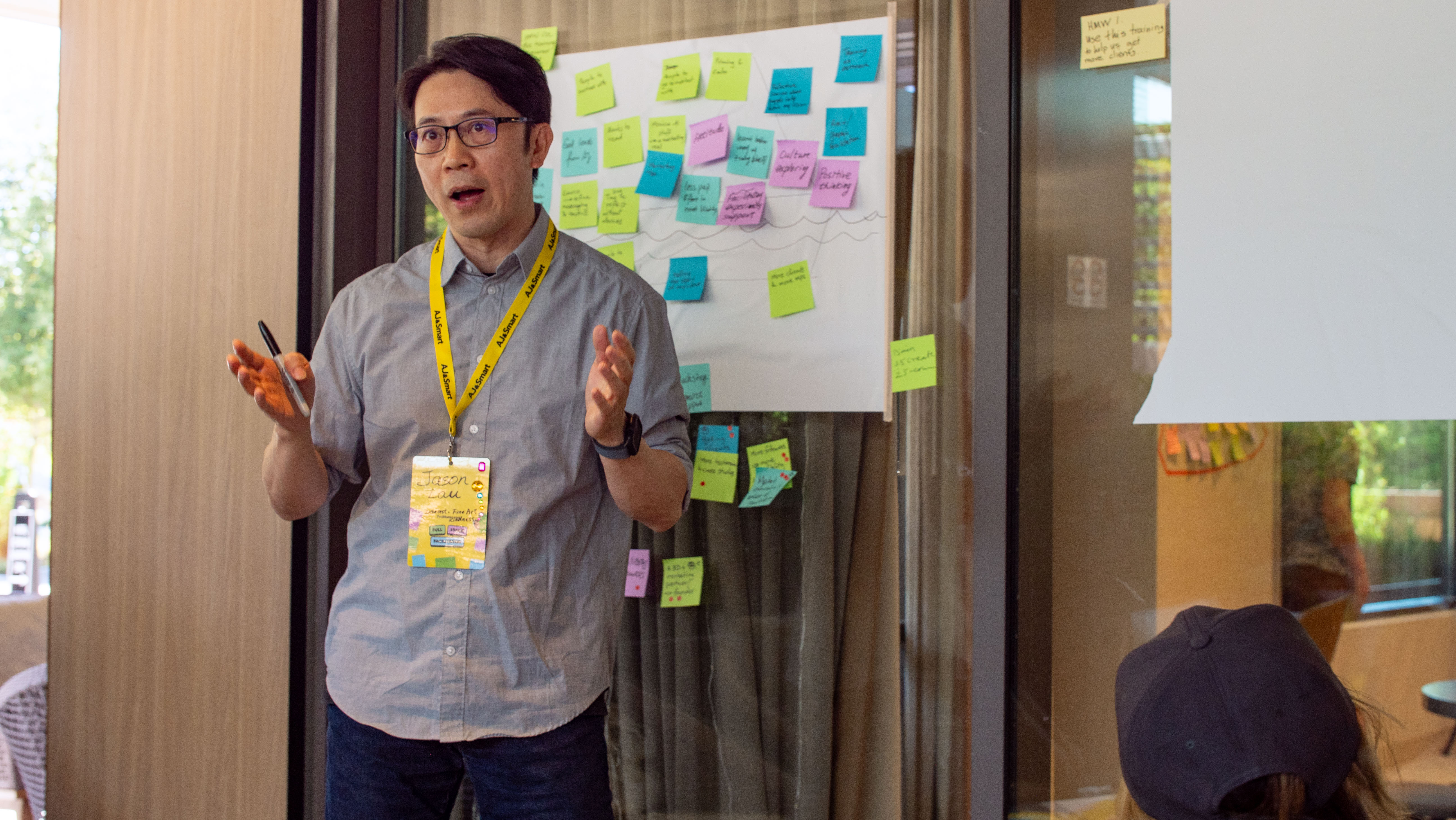 A facilitator wearing an AJ&Smart lanyard presents in front of a whiteboard covered in colorful sticky notes during a workshop session