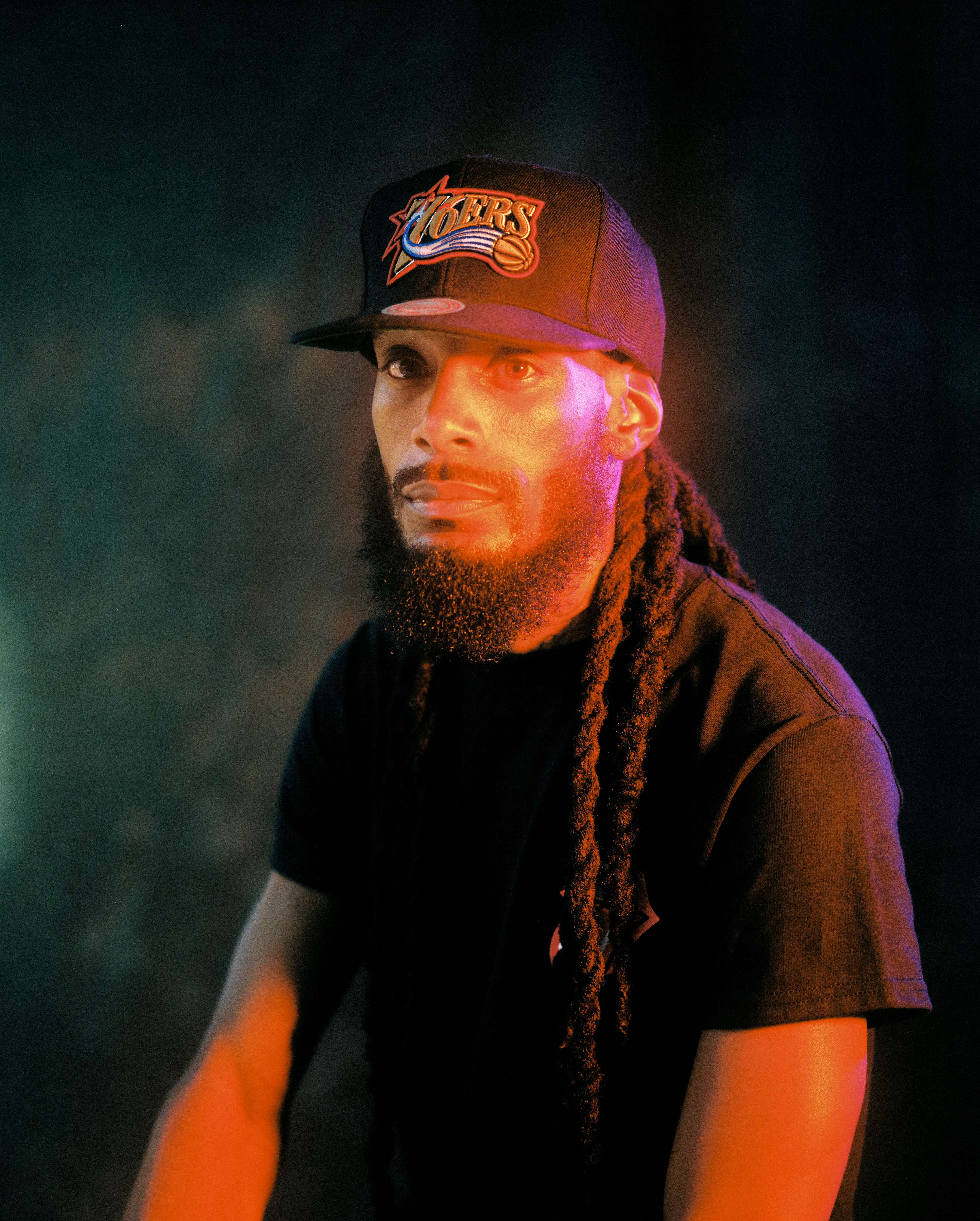 A portrait of Macnificent, a producer from the Crash Dummy collective, wearing a black T-shirt and a black 76ers cap. The image features dramatic lighting with warm orange and cool blue tones, highlighting his long dreadlocks and focused expression.