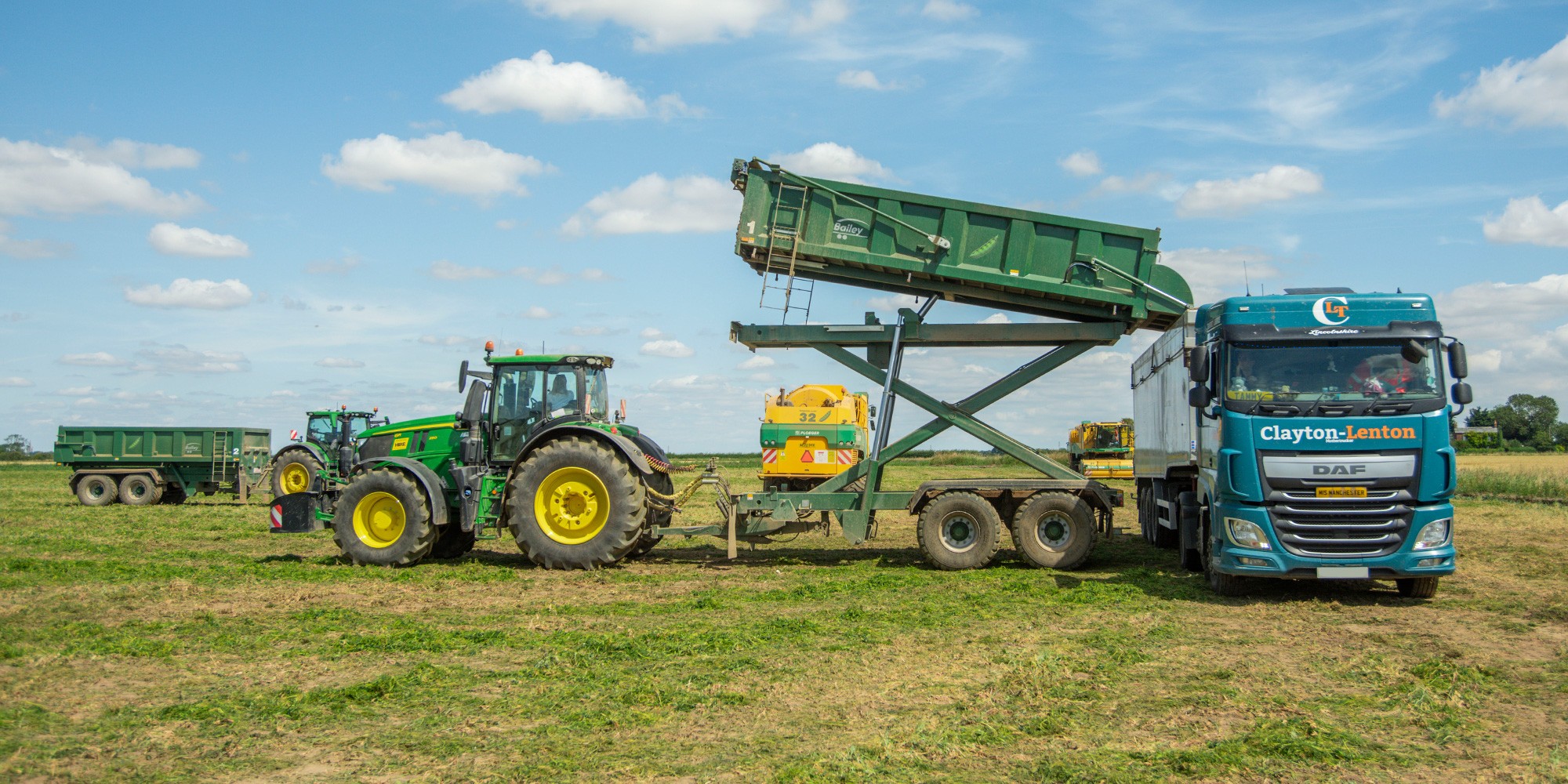 A John Deere tractor with a raised conveyor belt loading harvested peas into a Clayton-Lenton DAF truck in an open field, with additional farm trailers visible in the background under a blue sky with white clouds.