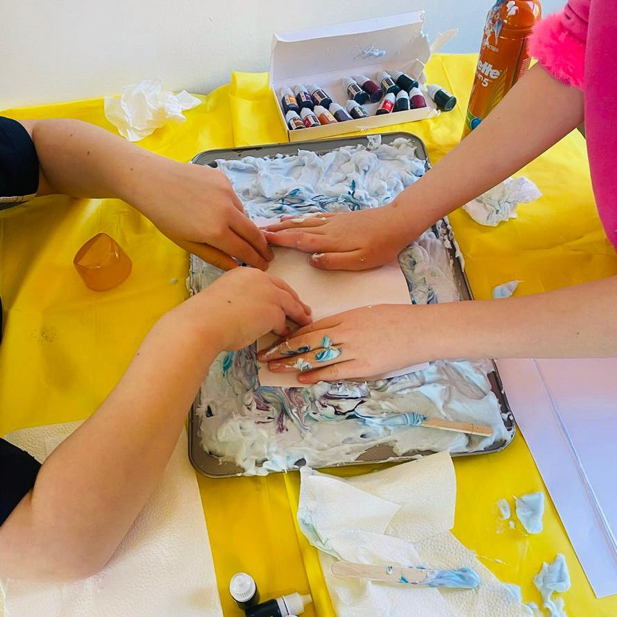 Two pairs of children's hands are pressing down on paper that's on top of paints to create a printed transfer