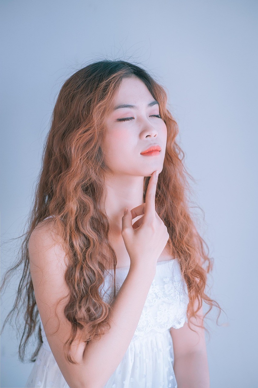 Close-up of a young woman with long, wavy auburn hair and closed eyes. Her right index finger gently touches her chin, and her lips are slightly parted, with a touch of coral lipstick. She wears a white, strapless dress with lace detailing. The background is a soft, muted blue.