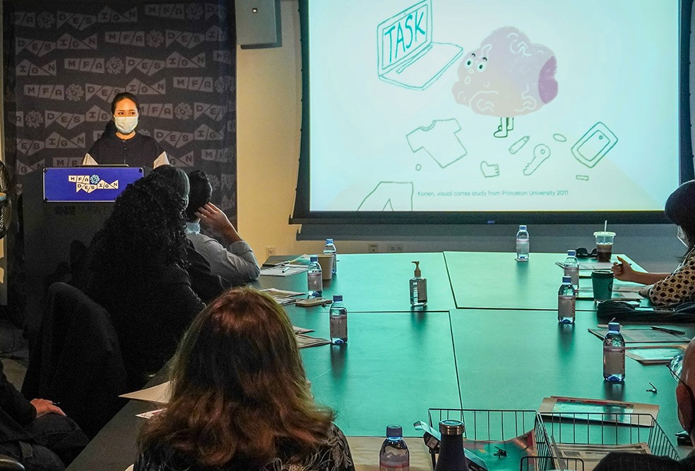 A woman is presenting from a stand in front of a few people sitting around the table, and she has an illustration projected on the right side on the wall.