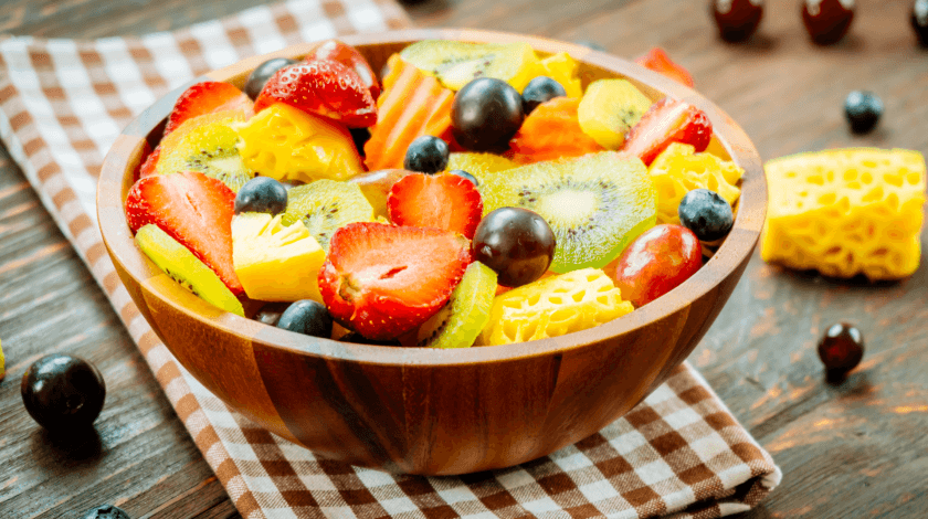 A wooden bowl filled with a vibrant assortment of fresh fruit, including sliced strawberries, kiwis, pineapples, grapes, and blueberries, rests on a checkered cloth atop a wooden table.