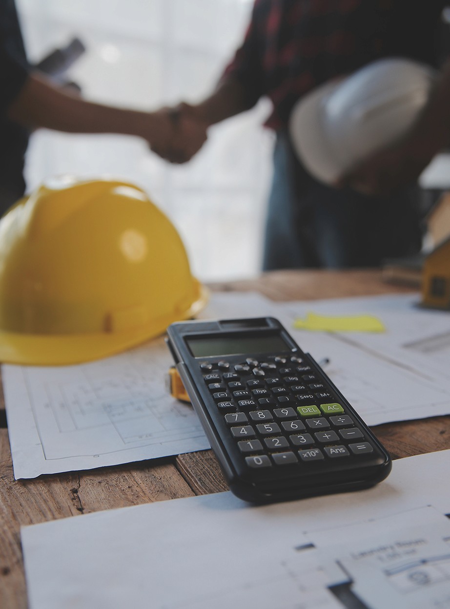 Close-up de uma calculadora sobre plantas de papel em uma mesa de madeira, com um capacete amarelo ao lado. Ao fundo, de forma desfocada, dois profissionais apertam as mãos. A imagem representa a complexidade de conciliar cálculos manuais com acordos de medição, um desafio que a Captura da Realidade digital ajuda a mitigar.