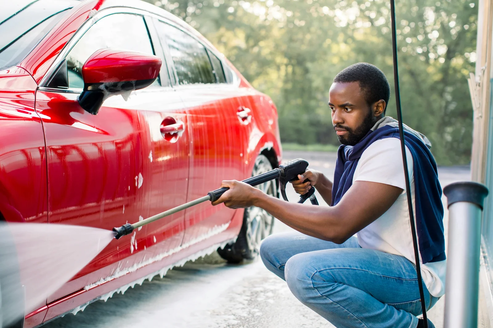 Man washing red car with pressure washer at outdoor carport during daytime.