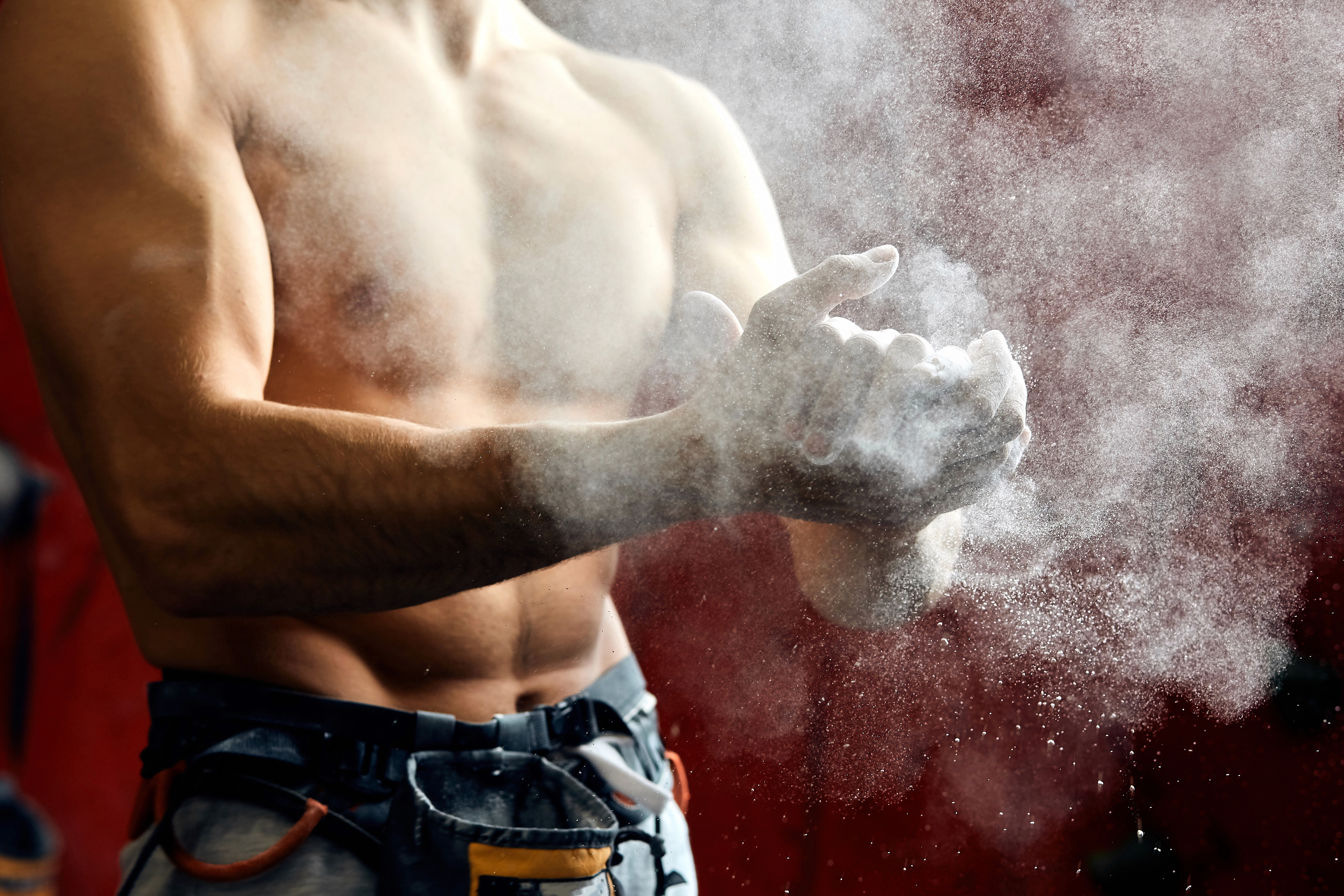 A person applying powdered chalk to their hands, likely in preparation for rock climbing or weightlifting. The cloud of chalk dust is visible in the air, while the person wears a harness around their waist, suggesting involvement in an athletic activity that requires grip and strength. 