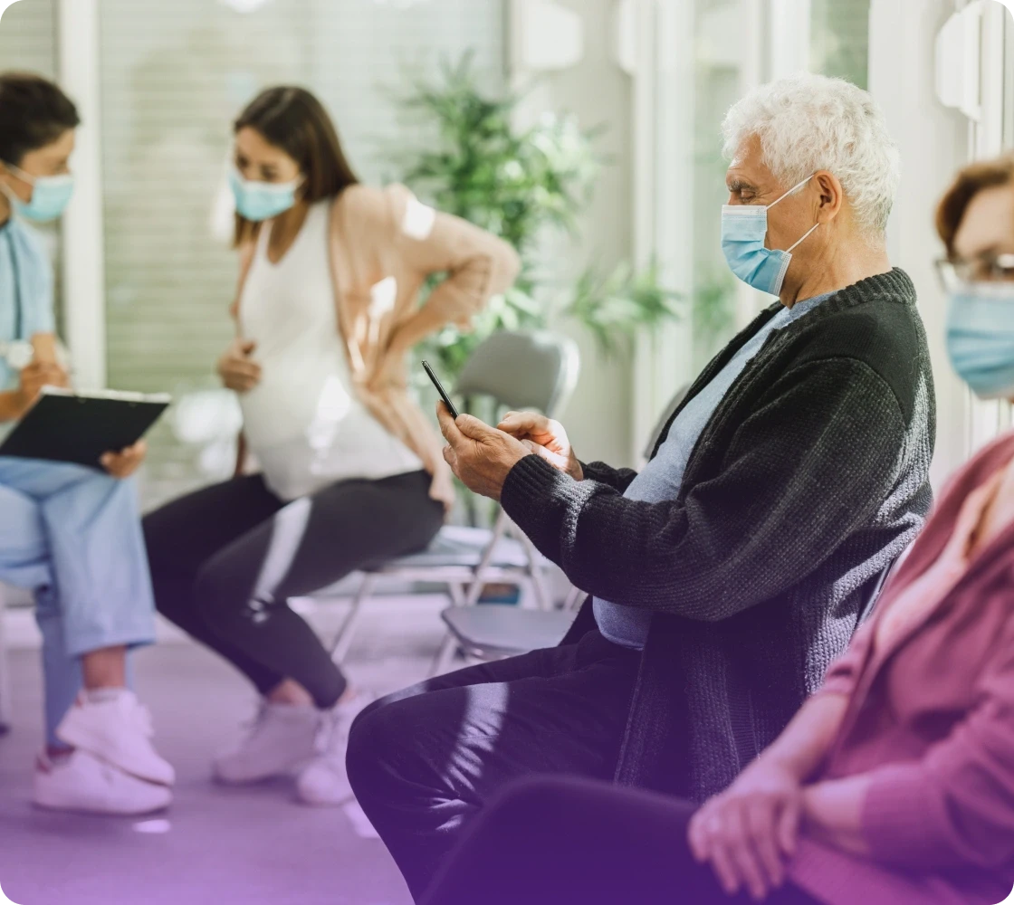 Four people wearing masks are sitting in a waiting area, engaged in conversation or using devices.