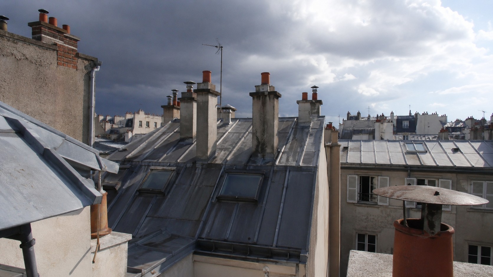 Toiture parisienne en zinc sous un ciel d’orage d’été, avec cheminées, velux et gouttières exposés aux risques d’infiltration après fortes pluies.