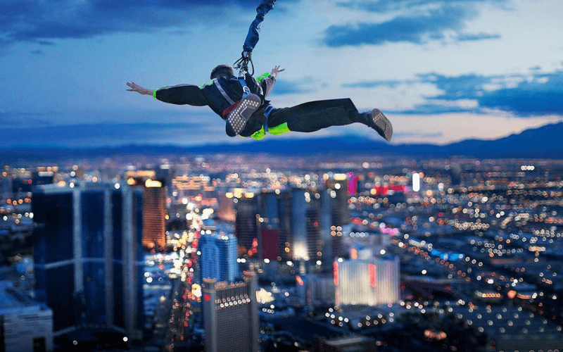 A SkyJump participant hangs mid-air, arms and legs outstretched with the lights of the Vegas Strip below.