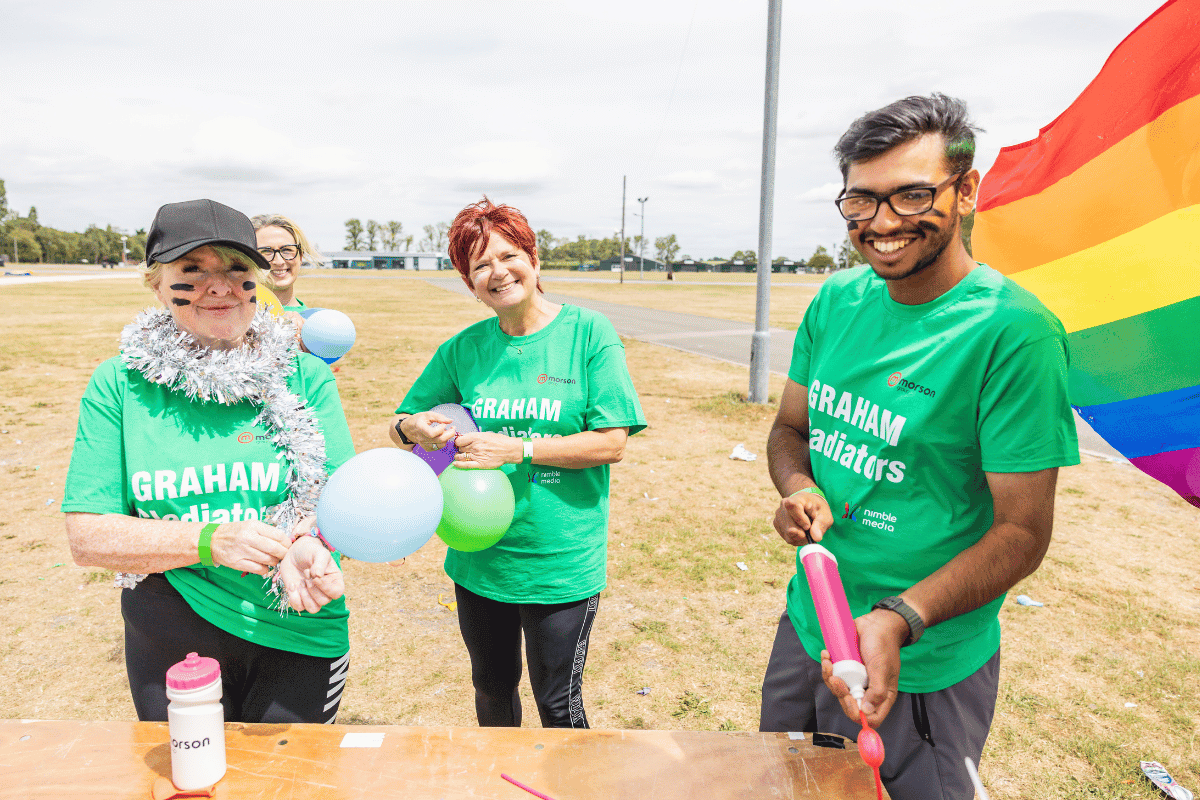 Outdoor team-building event with participants wearing matching blue shirts, taking part in inflatable obstacle courses and games, smiling and cheering, with the words 'ENTER A TEAM' in large bold white and green text across the image.