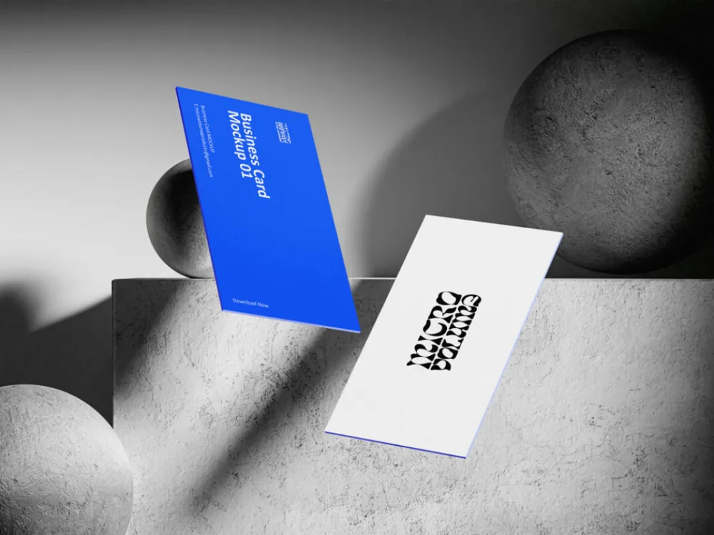 Floating business card mockup showcasing two designs: one in vibrant blue with white text and another in white with a bold black logo. Set against a textured concrete surface with spherical objects and dramatic grayscale lighting.